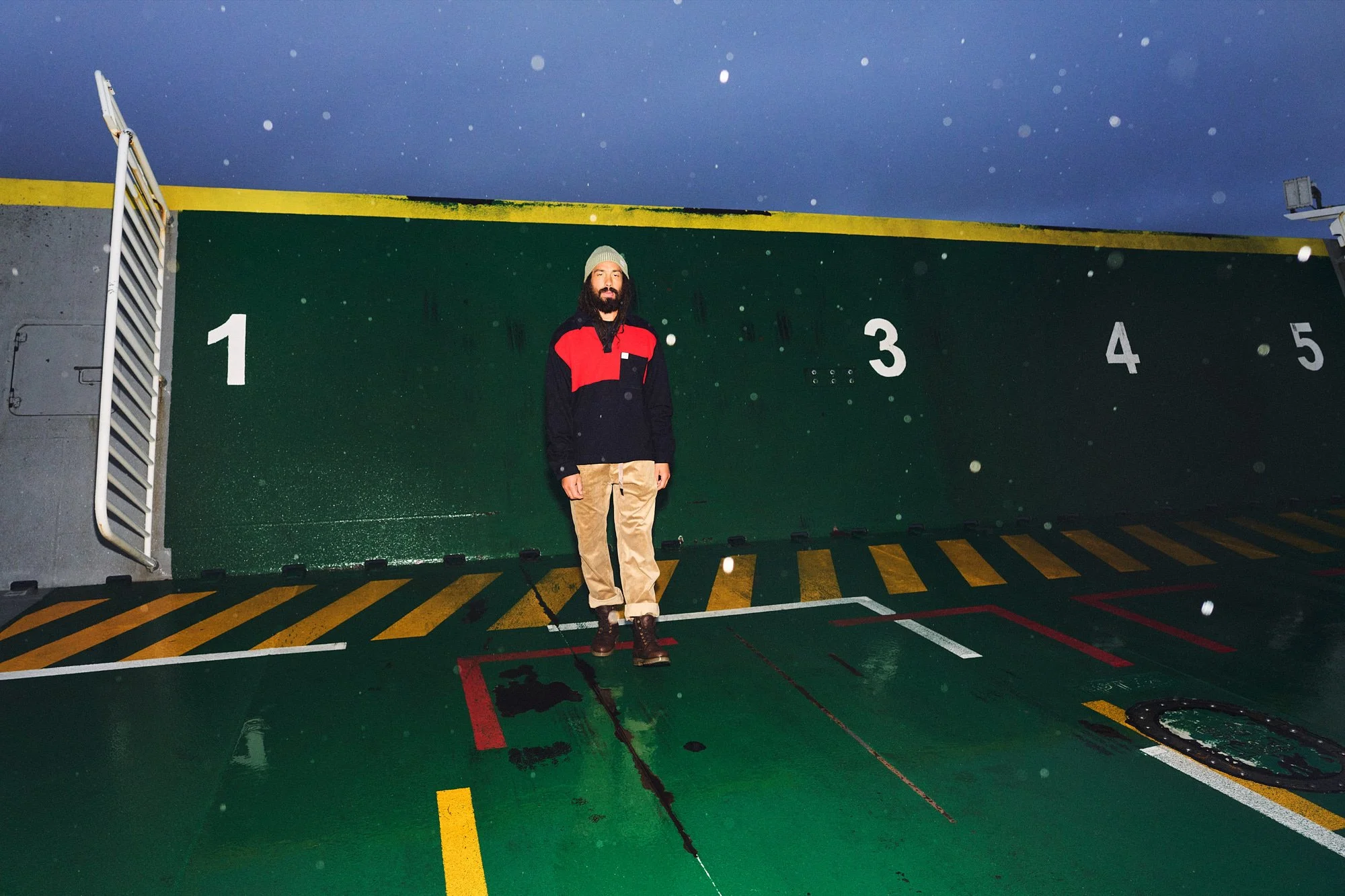 A man standing on the deck of a boat at night. The deck has yellow and black striped markings, and the background includes a green wall with white numbered markings from 1 to 5. The sky is dark and starry, with raindrops visible in the air.