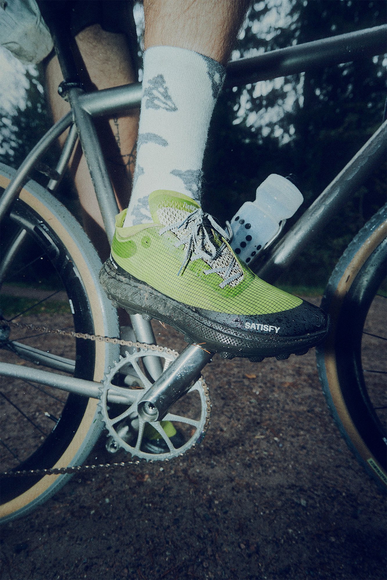 Close-up of a person's foot on a bicycle pedal, wearing a bright yellow and black cycling shoe, water droplets on the shoe, and a white sock with gray runes or designs. The bicycle has a silver crankset and black frame, and the background shows a mud