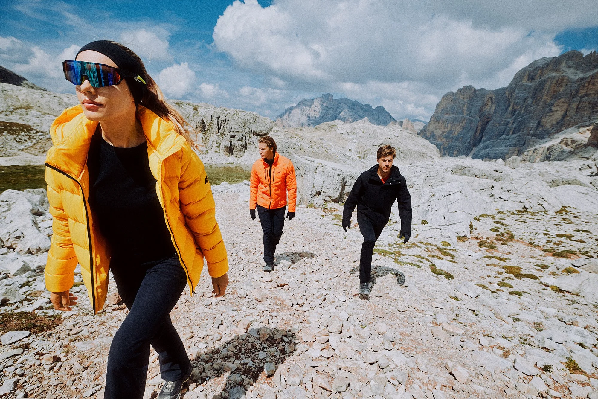 Three people hiking on a rocky mountain trail under a partly cloudy sky. One woman in the foreground is wearing a yellow jacket and sunglasses, leading the group. Two men are behind her, one in an orange jacket and the other in black.