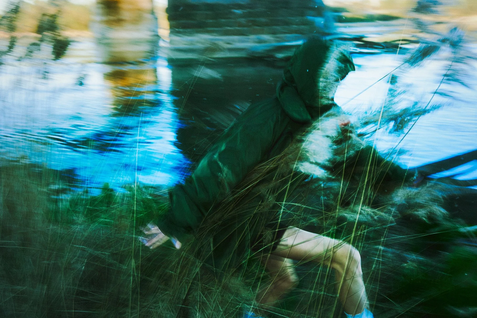 A person in a black hoodie and beige shorts crouches near a body of water, with a fishing rod in hand, surrounded by tall green grass and trees reflected in the water.