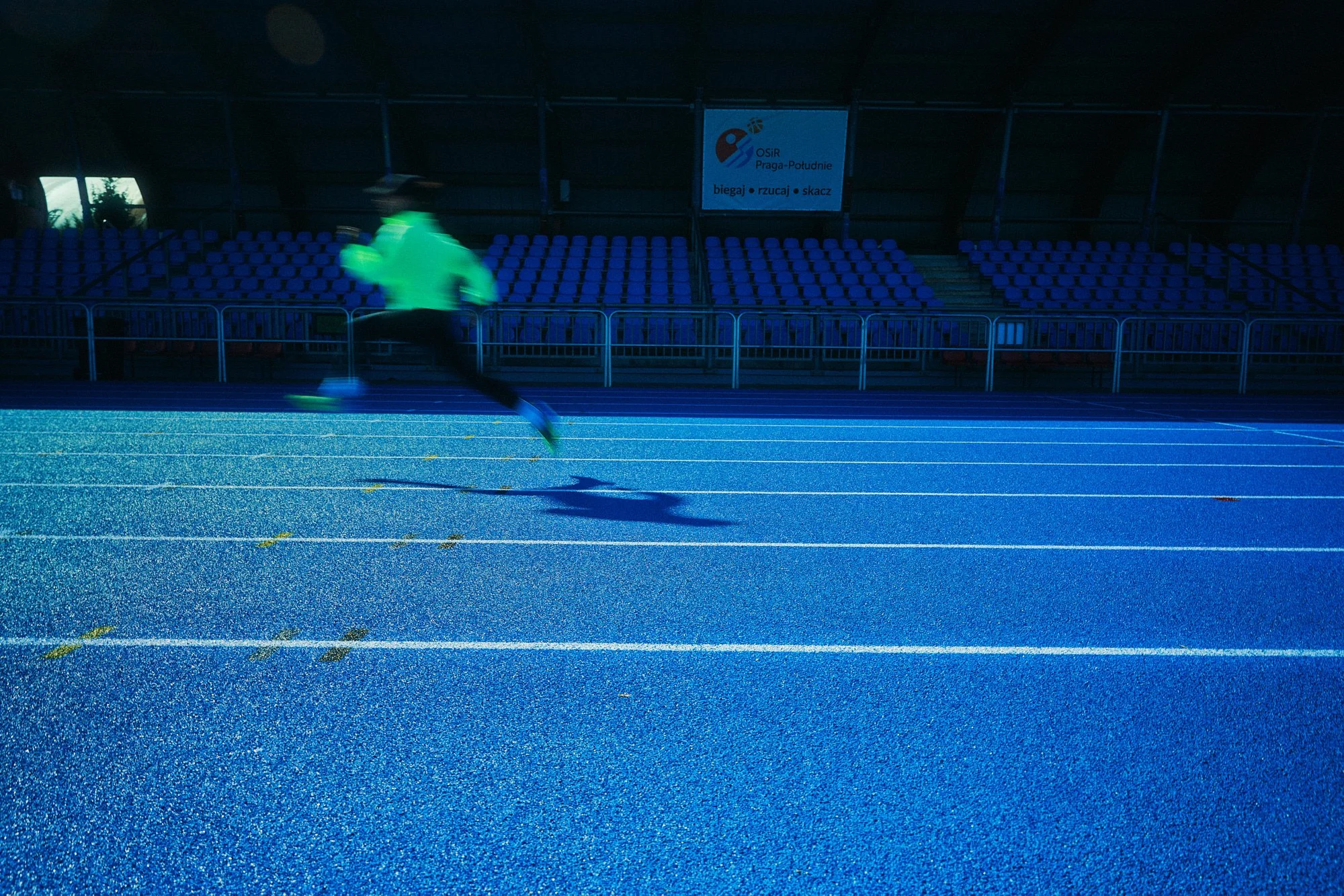 A person running on a blue outdoor track at night, wearing a neon green jacket and black pants, with empty blue stadium seats in the background.