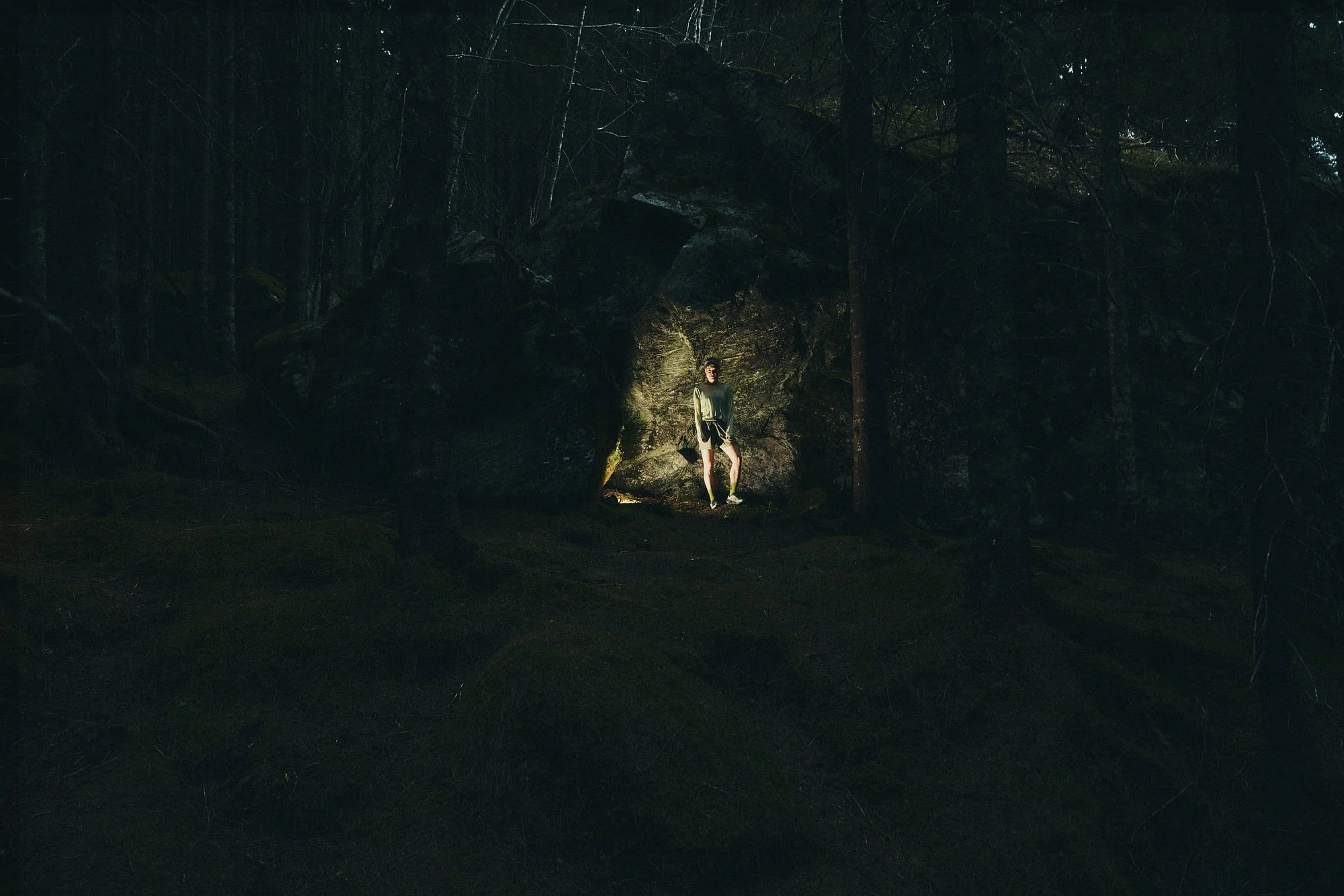 A person standing in a forest clearing illuminated by a light source, surrounded by trees and large rocks.