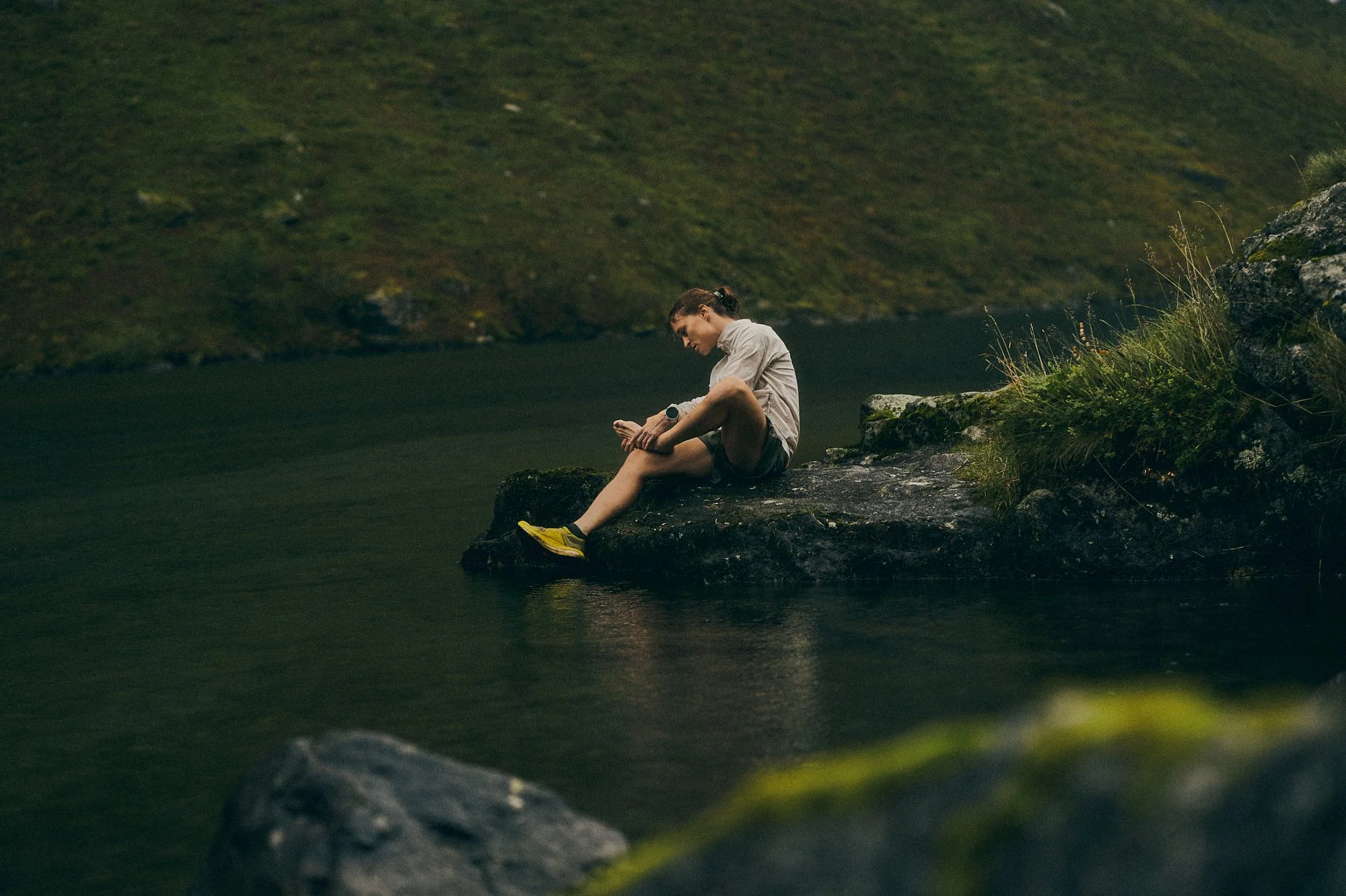 Woman sitting on a rock by a calm river, looking at her watch in a lush, green landscape.