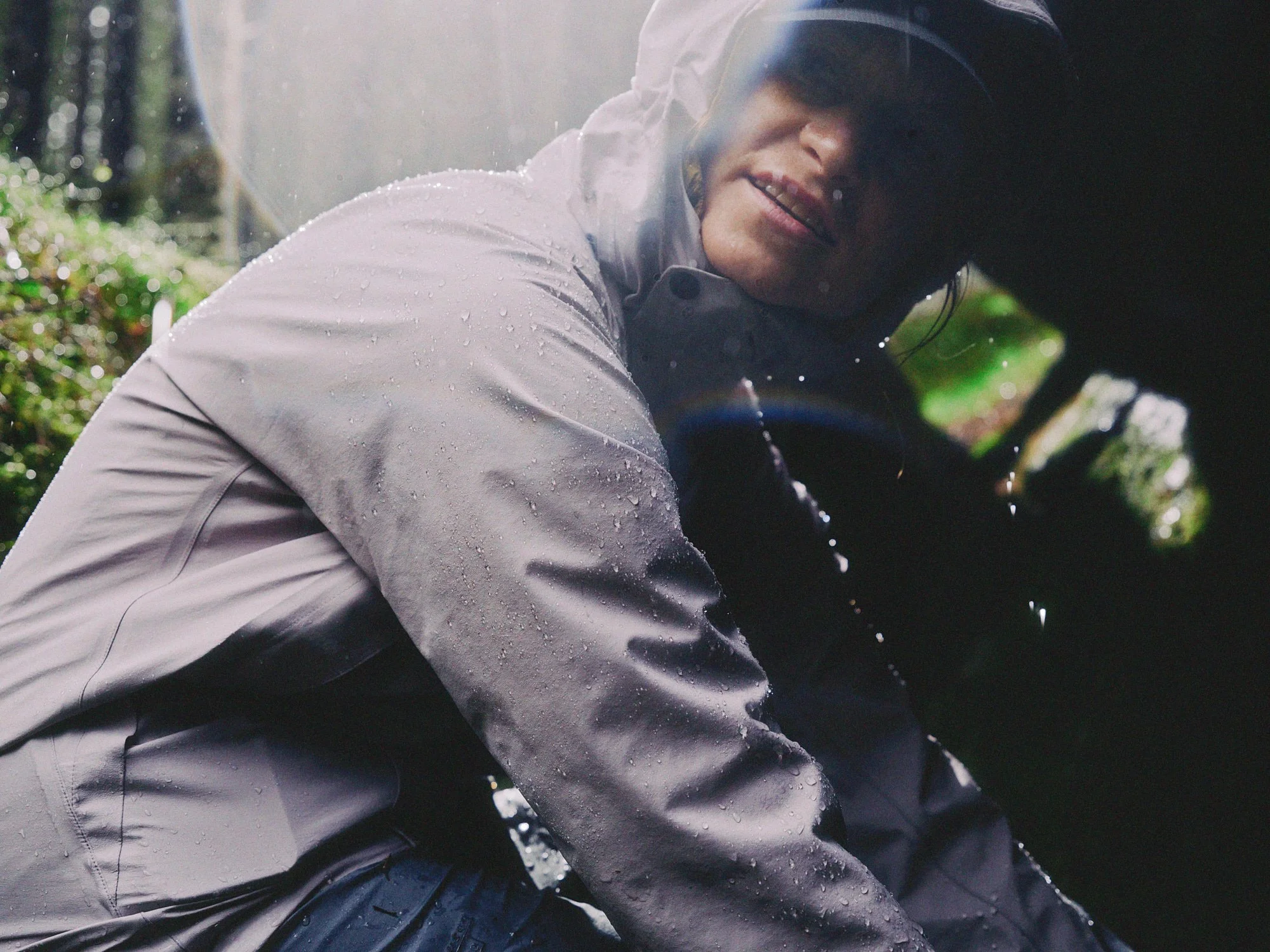 A woman in a rain jacket and cap crouching outdoors, with water droplets on her jacket, in a forest setting.