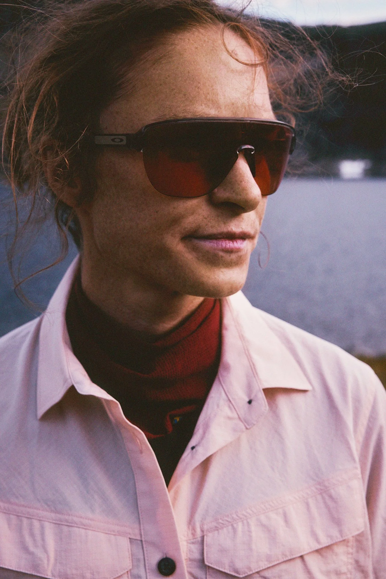 Close-up of a woman with wavy red hair, wearing large sunglasses, a light pink jacket, and a dark red turtleneck, standing outdoors near a body of water.