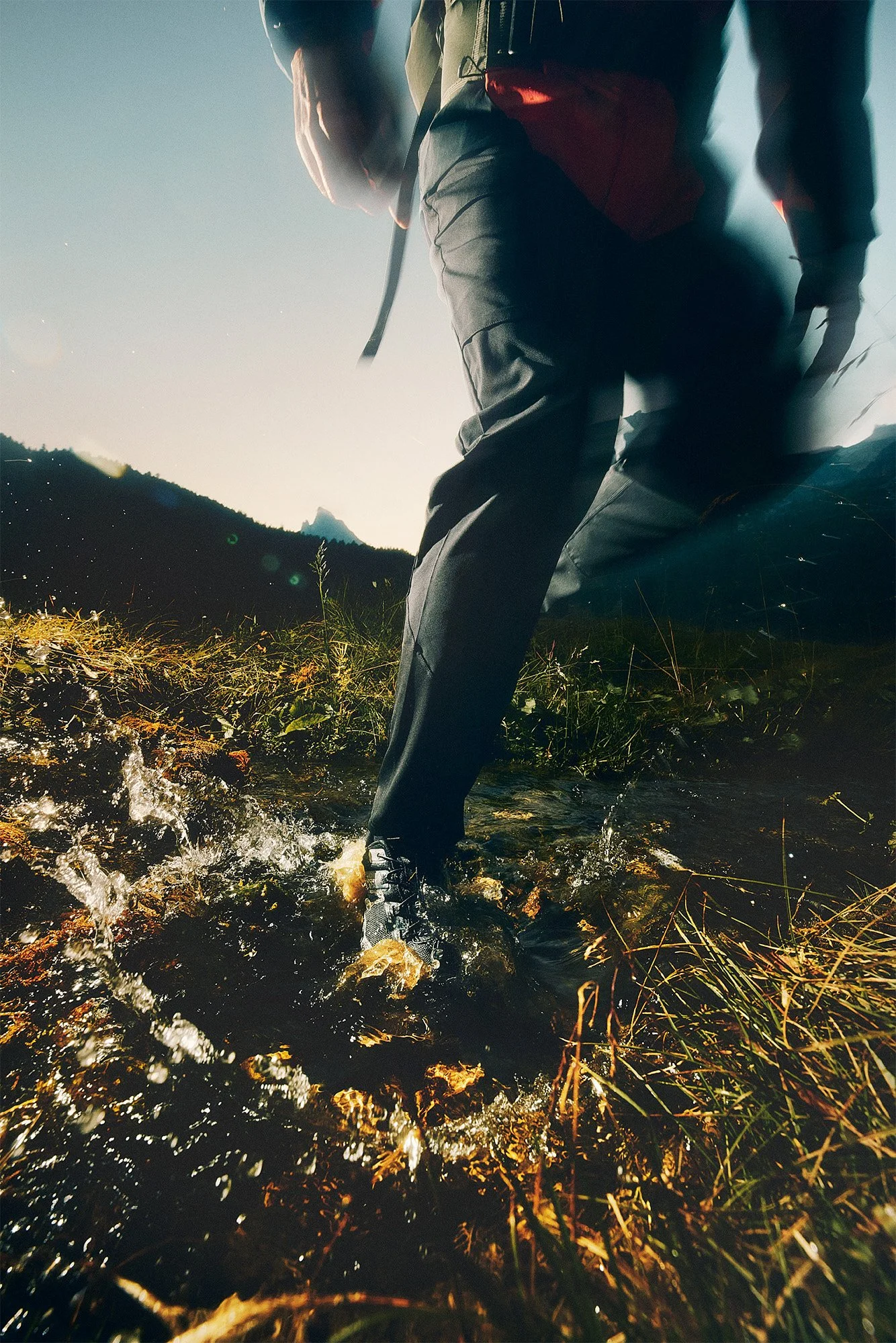 Hiker crossing a stream in a mountainous landscape at sunset.
