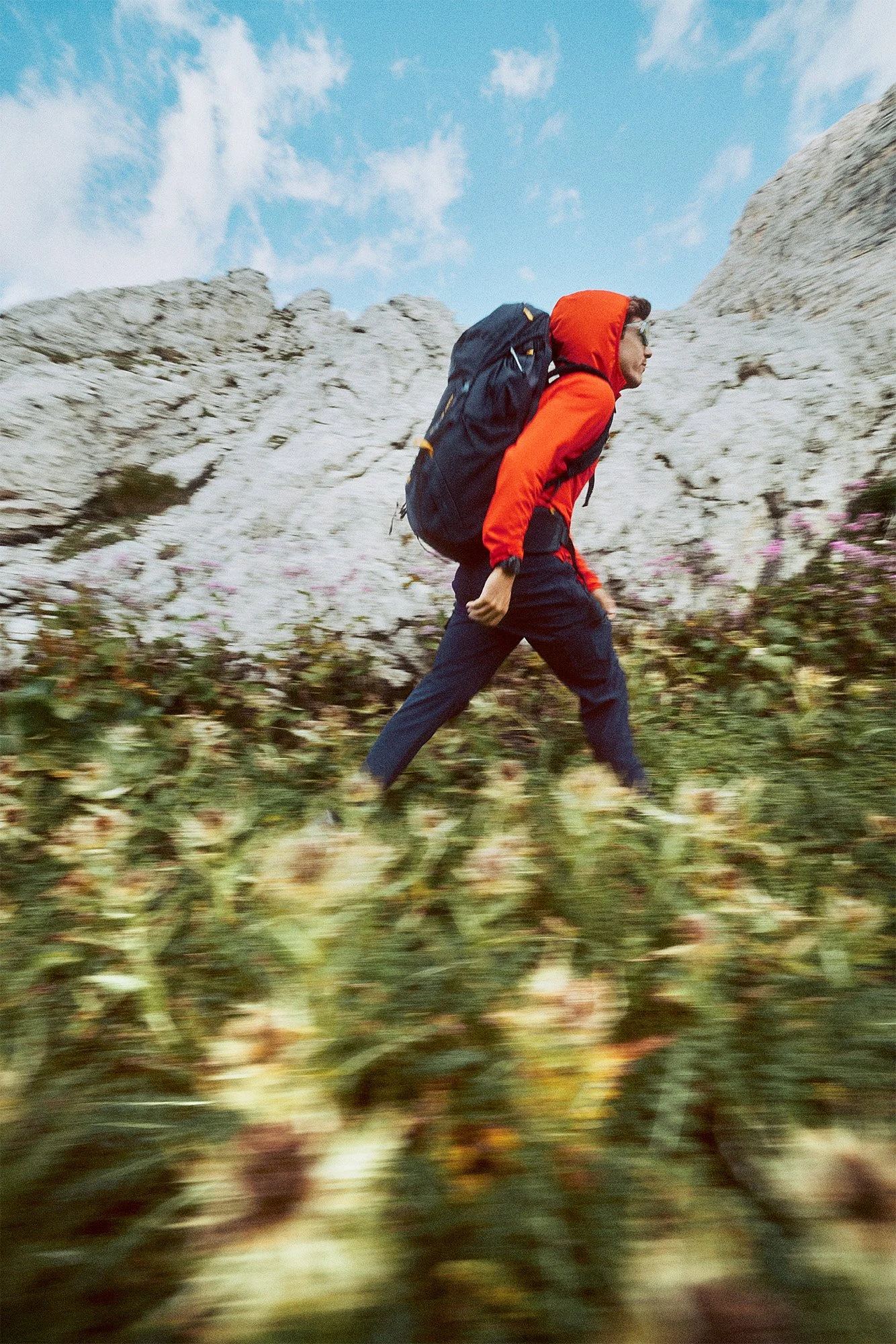 Person hiking in outdoor mountainous landscape with backpack, wearing red jacket and dark pants.