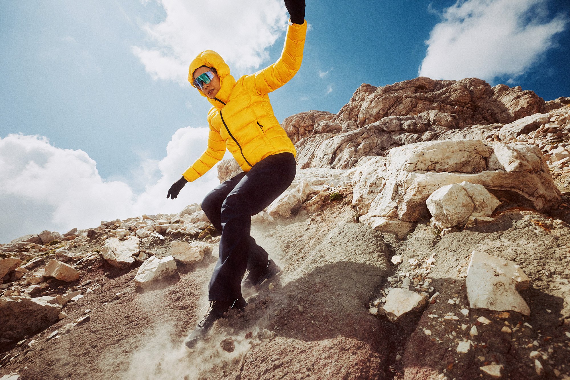 A person in a yellow jacket, black pants, gloves, and sunglasses snowboarding down a rocky mountain slope under a cloudy sky.