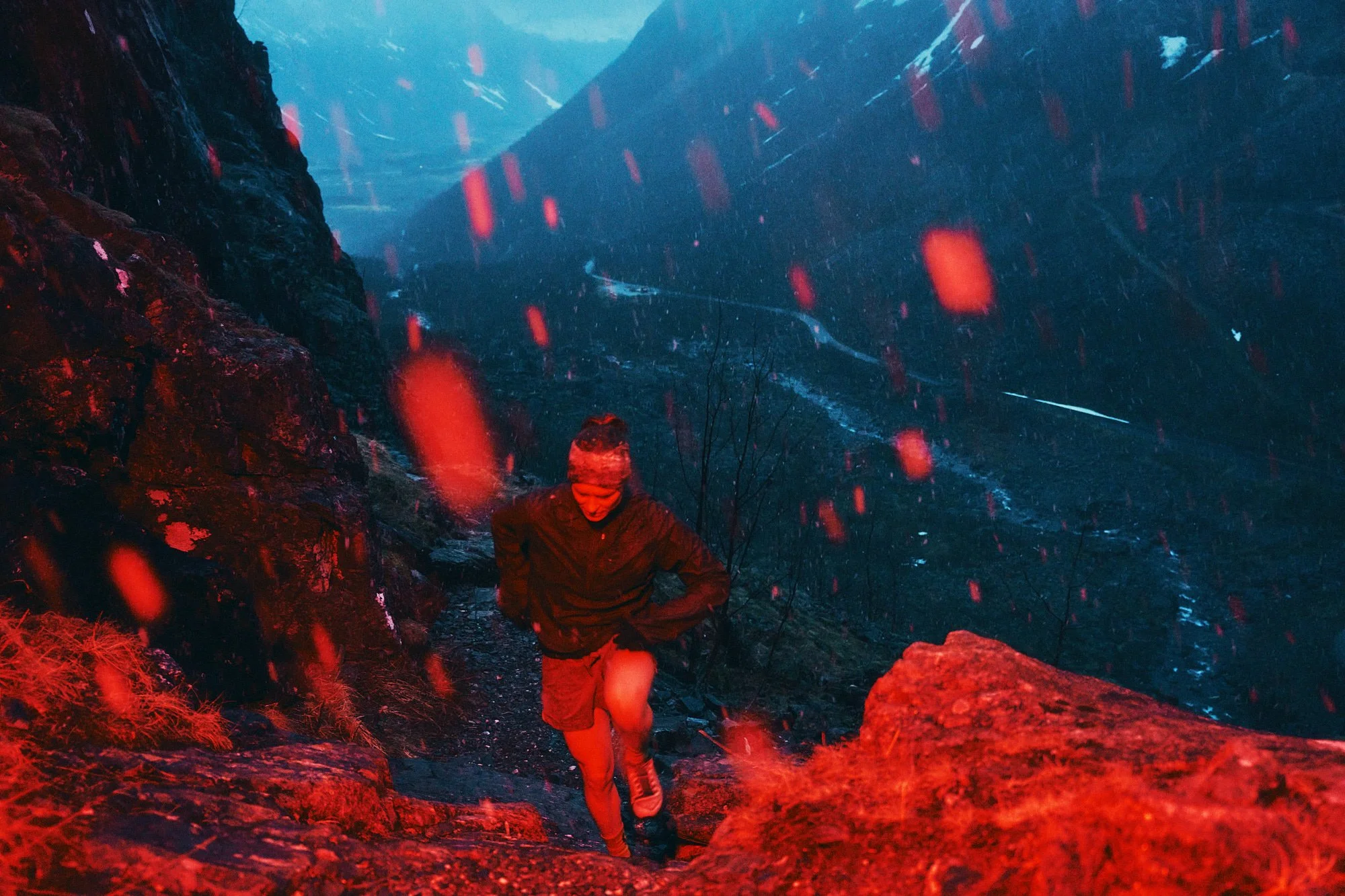A person hiking on a trail in a mountainous area during a rainy night, illuminated with red lighting.