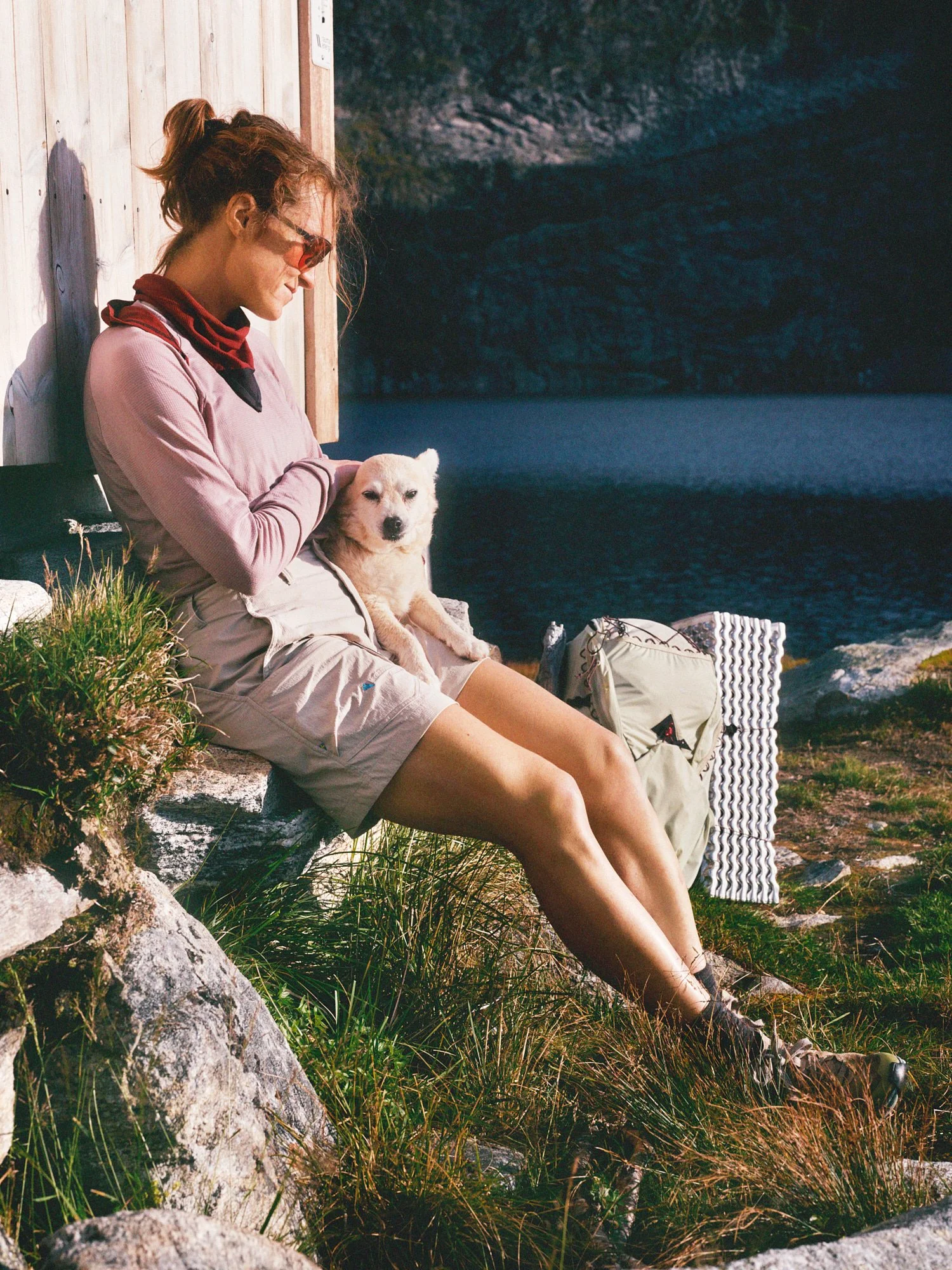 A woman sitting on a rock by a lake, holding a small dog in her lap, with a backpack and foam pad nearby, wearing outdoor clothing and sunglasses, with a rocky cliff in the background.
