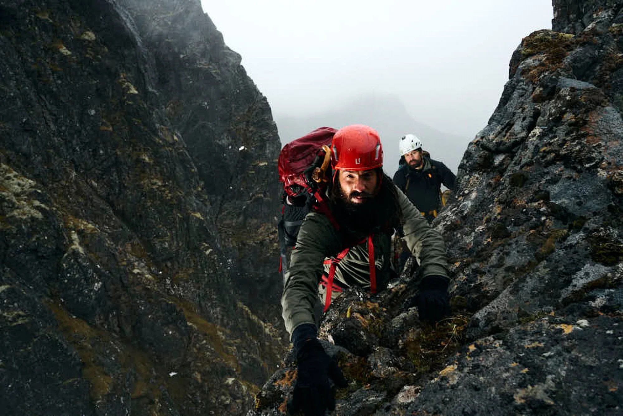 Two climbers ascending a steep rocky mountain on a foggy day, wearing helmets and carrying gear.
