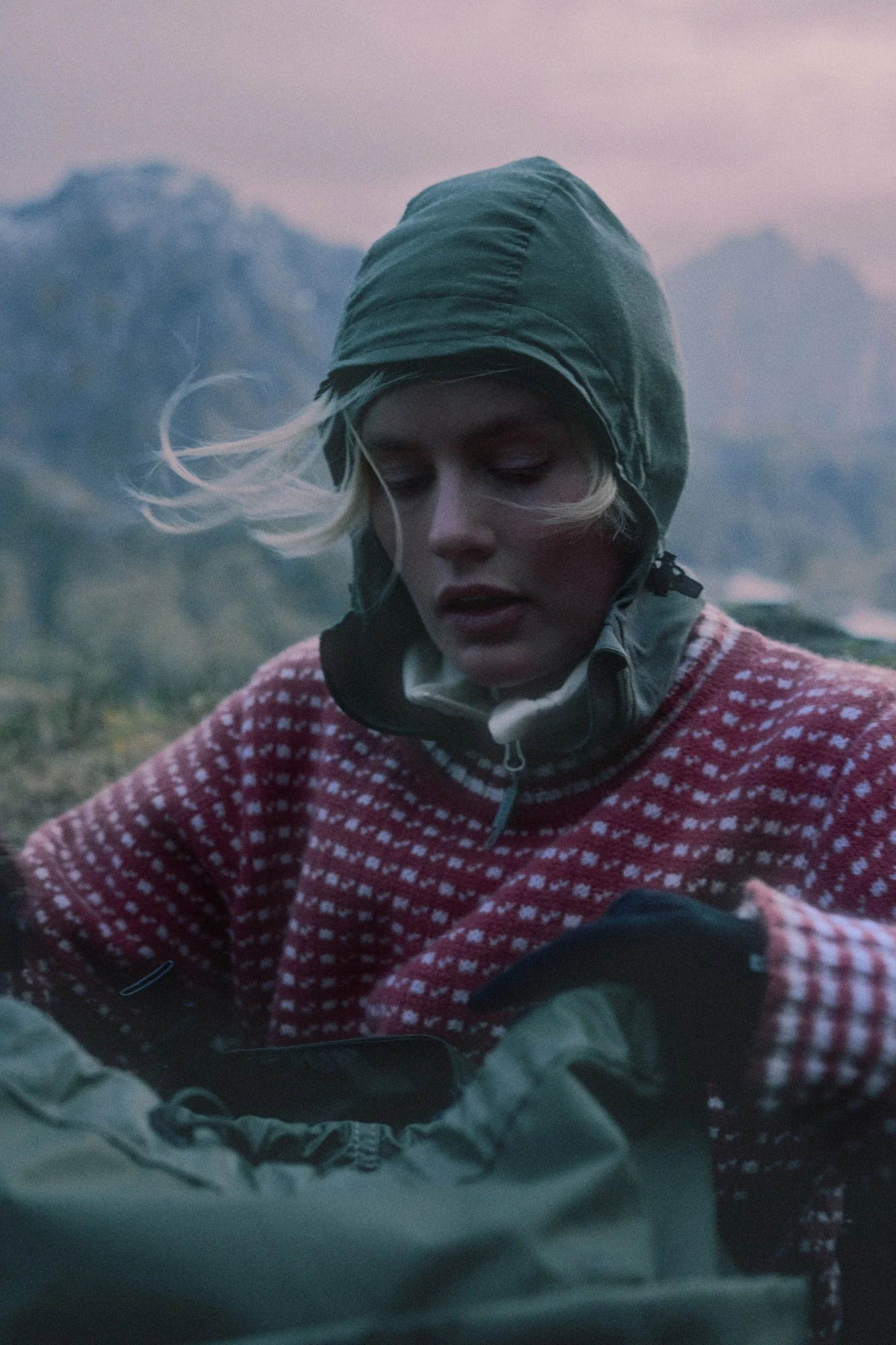 A woman dressed in outdoor gear, including a hoodie and patterned sweater, looking down in an outdoor mountainous landscape at dusk or dawn.