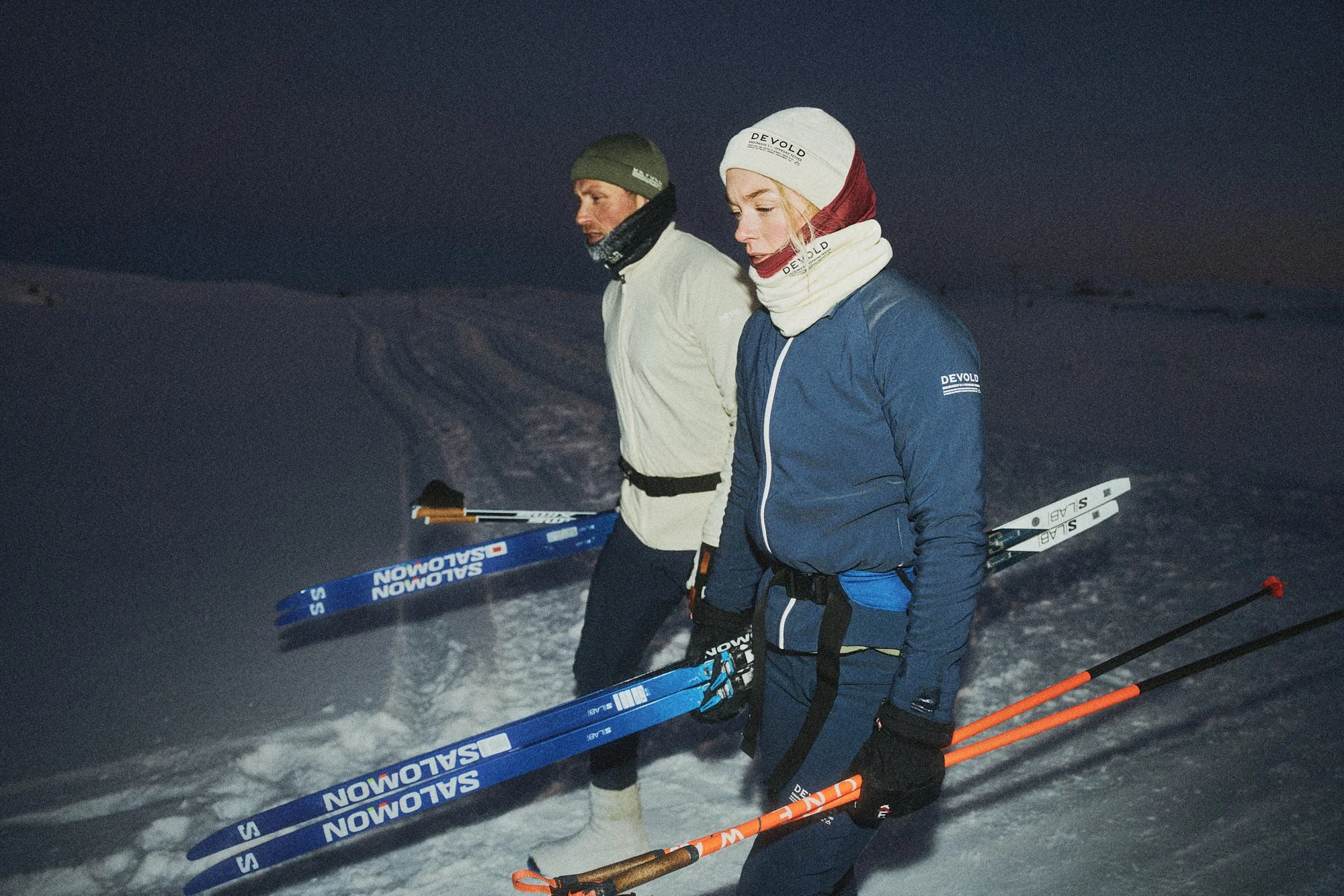 Two women in winter gear and holding cross-country skis walk through snow at night.