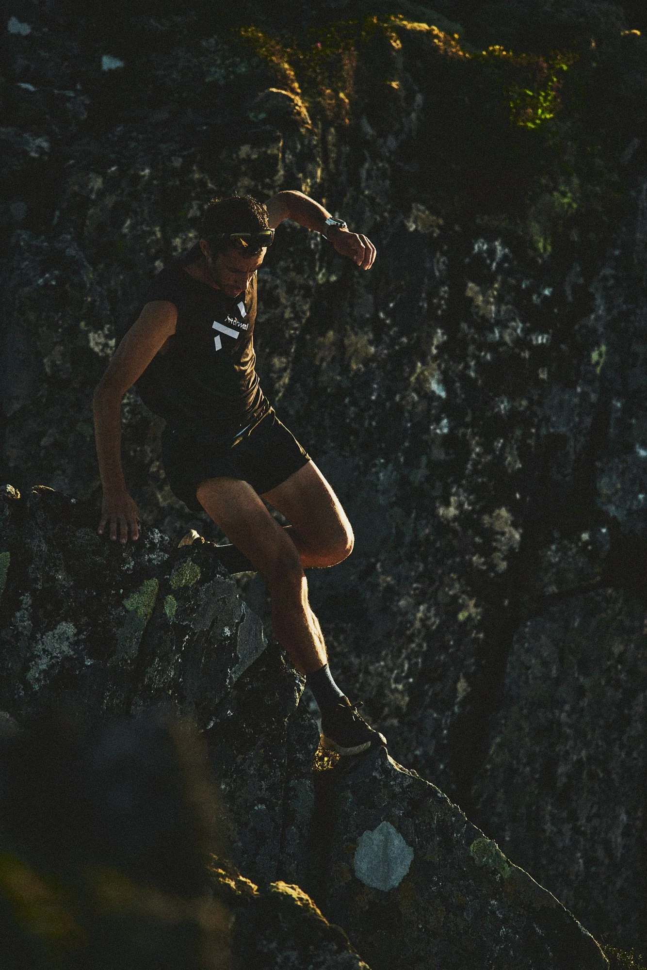 A person in black athletic clothing and sunglasses jumps down a rocky cliff at sunset.