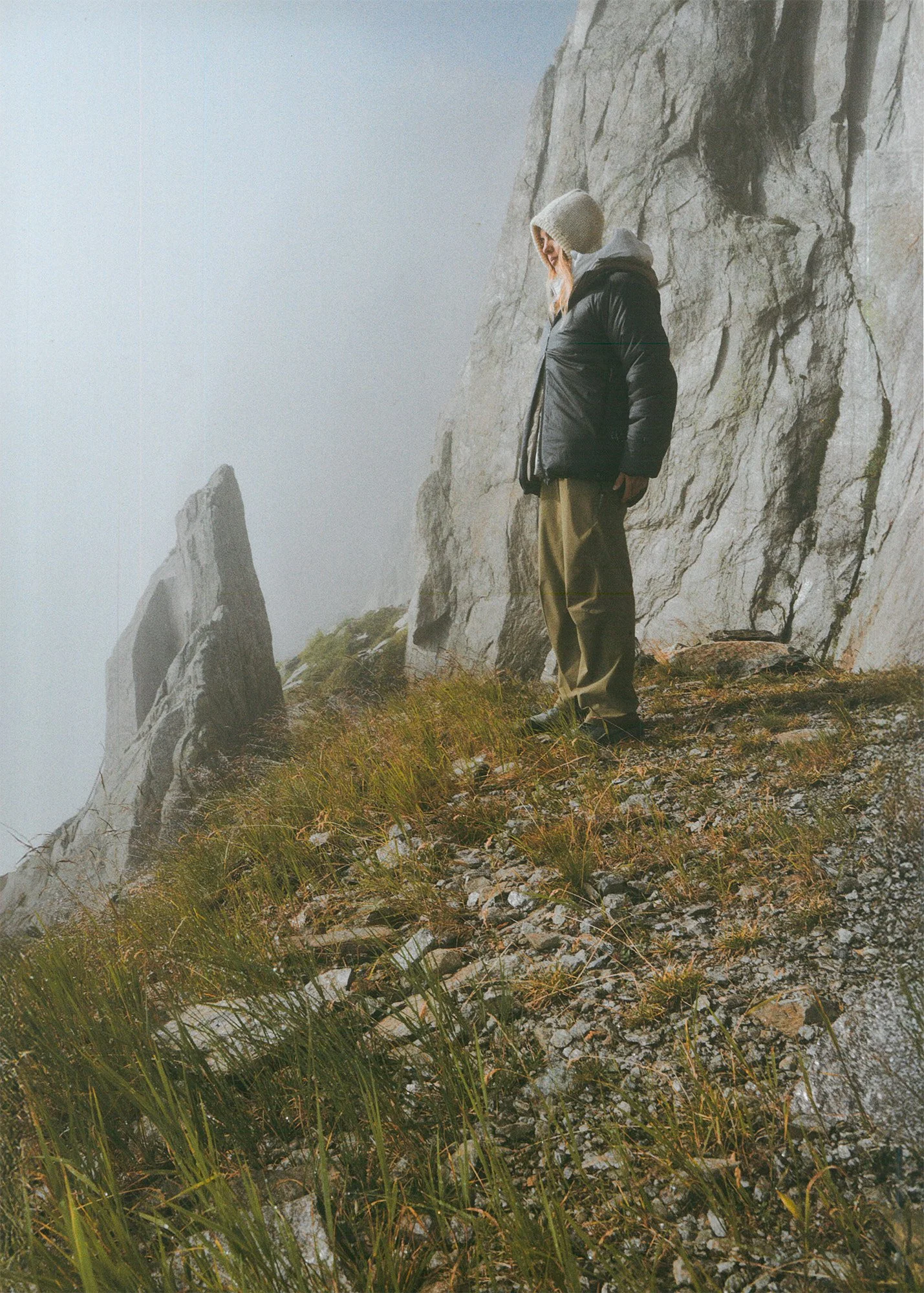 A person in a hoodie, black jacket, and khaki pants standing on a foggy, rocky mountainside