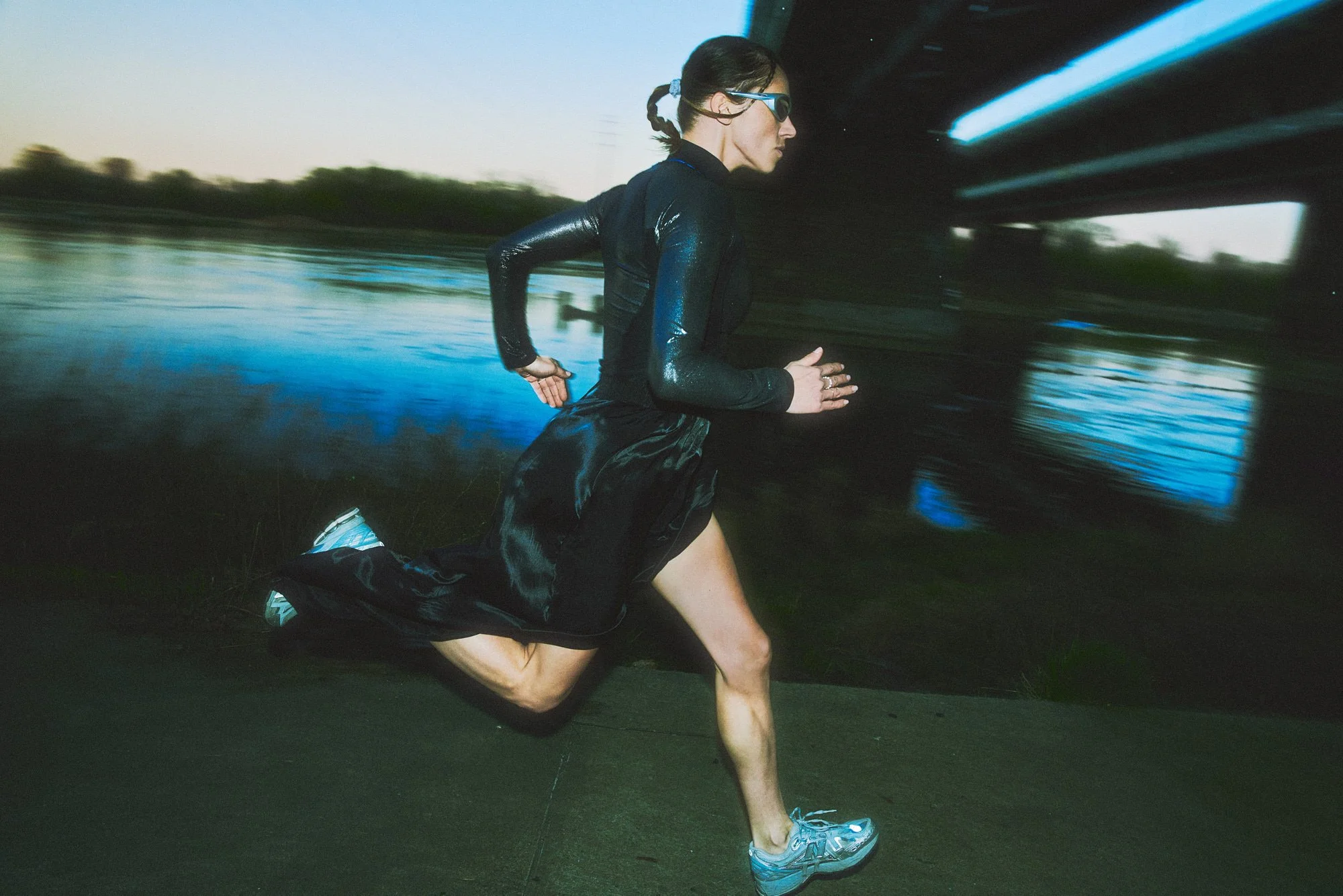 A woman wearing sunglasses and athletic running clothes is running outdoors near a body of water during twilight.
