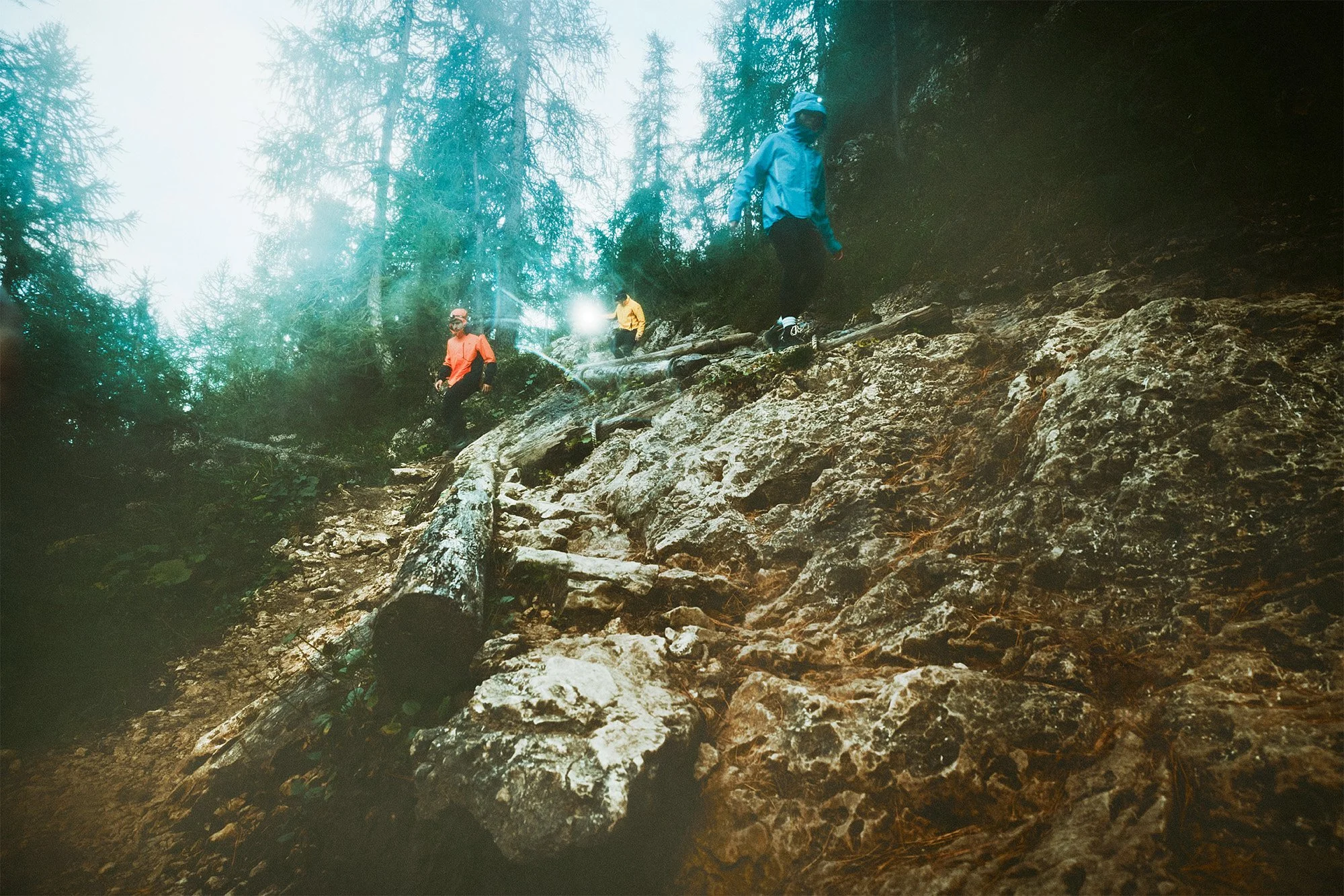 Three hikers climbing a rocky forest trail on a mountain, wearing colorful jackets and helmets.