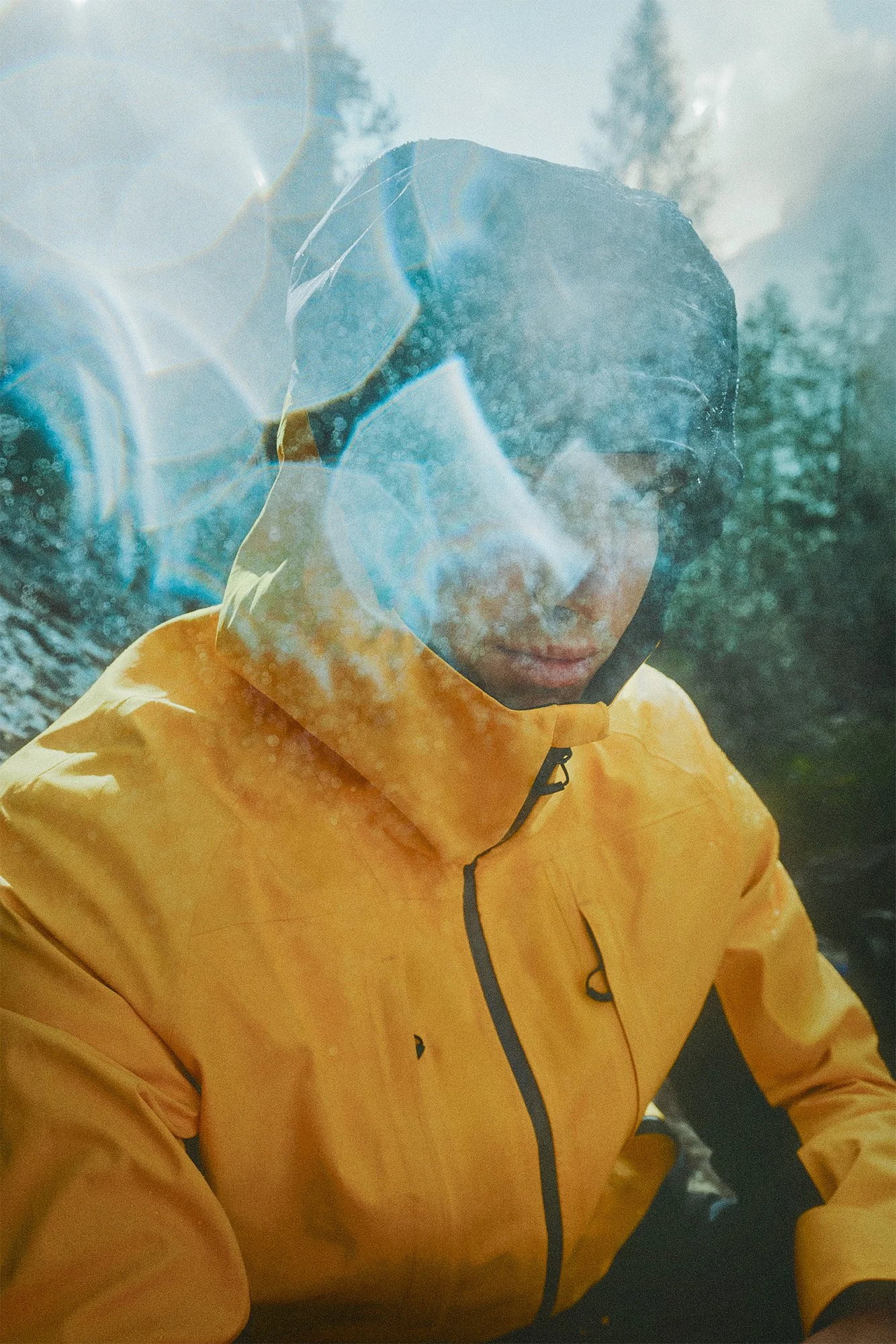 A person wearing a yellow jacket and a dark hood, looking down, with reflections of trees and clouds on the glass in front of them.