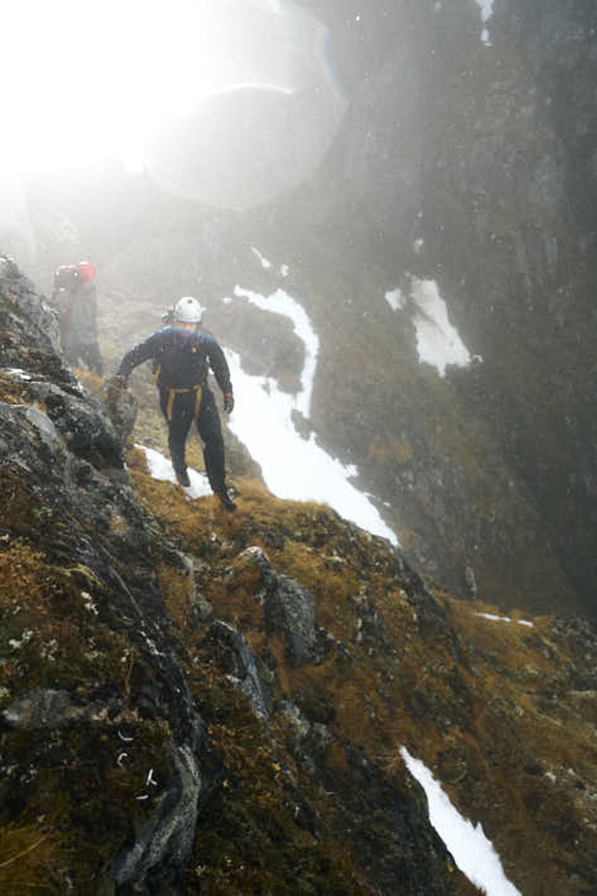 Two hikers with helmets and backpacks walking along a narrow, rocky trail on a mountain slope, with snow patches and a foggy atmosphere.