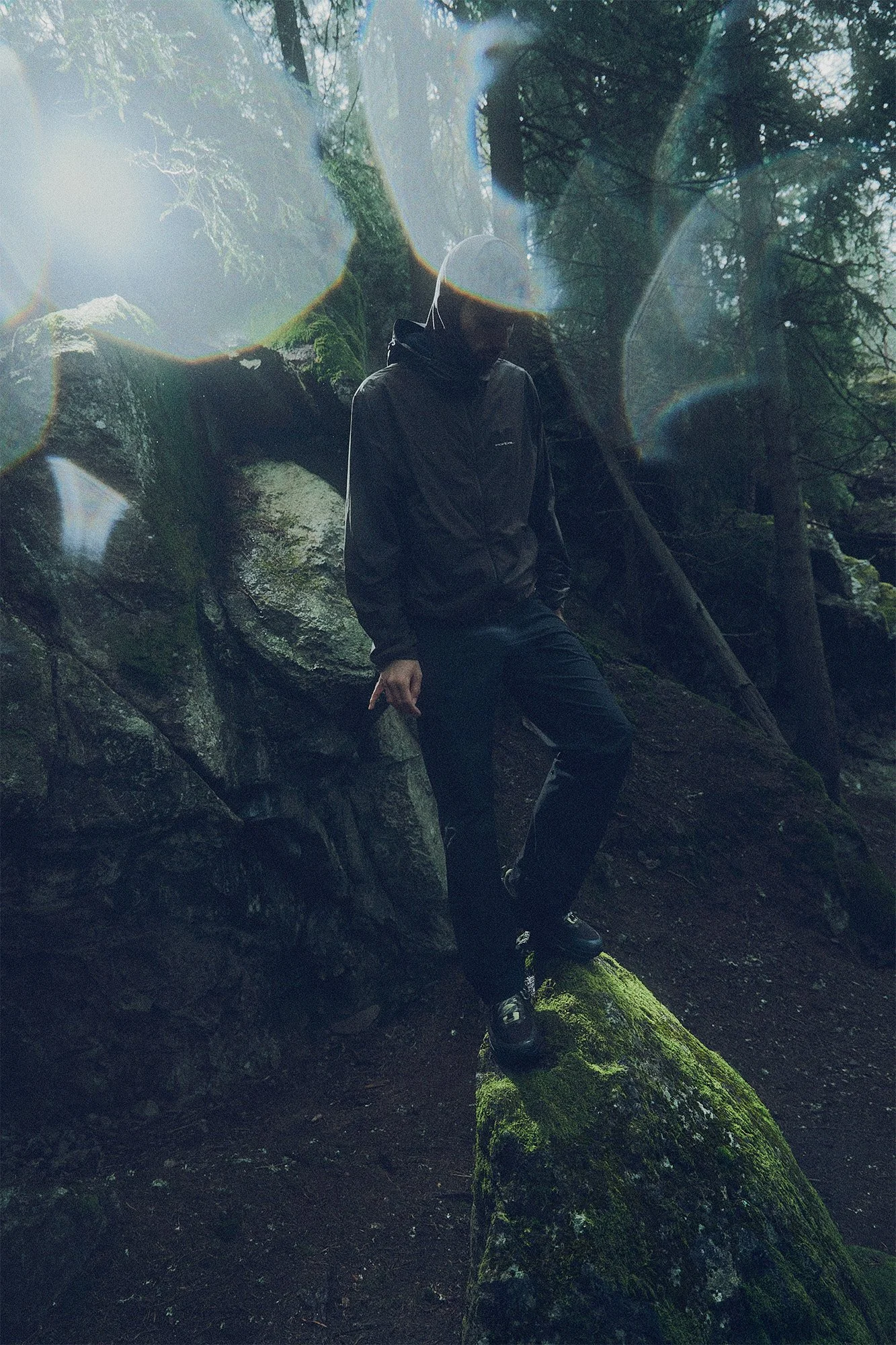 Person in black outdoor clothing standing on moss-covered rock in a forest, with sunlight filtering through trees.