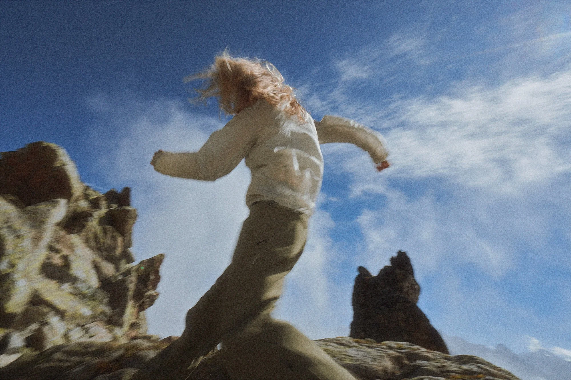 Person with long hair running or jumping on rocky terrain under blue sky with clouds