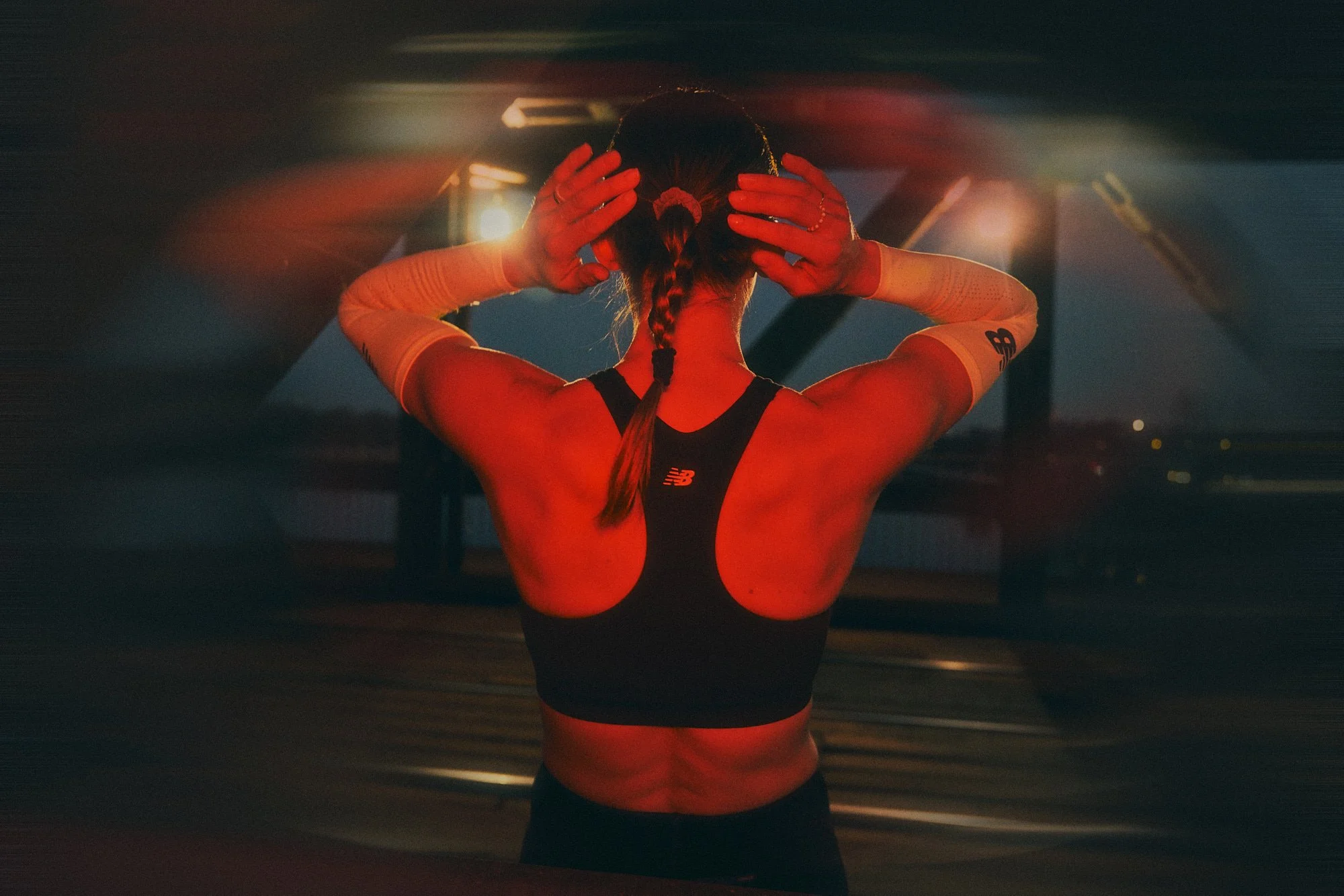 Back view of a female athlete adjusting her braided hair in a dimly lit gym, wearing a black sports bra and white arm sleeves.