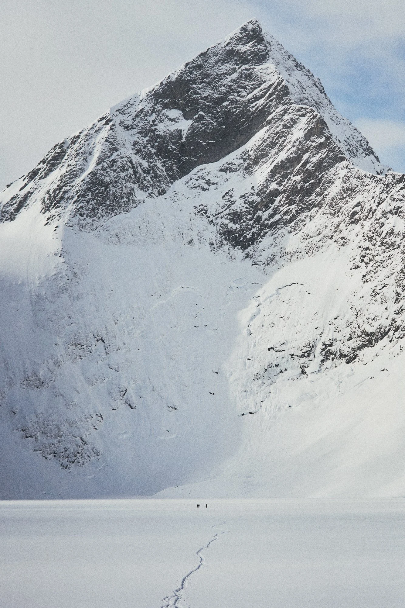 Snow-covered mountain with steep slopes and a peak, with two small figures and a trail of footprints in the snow in the foreground.