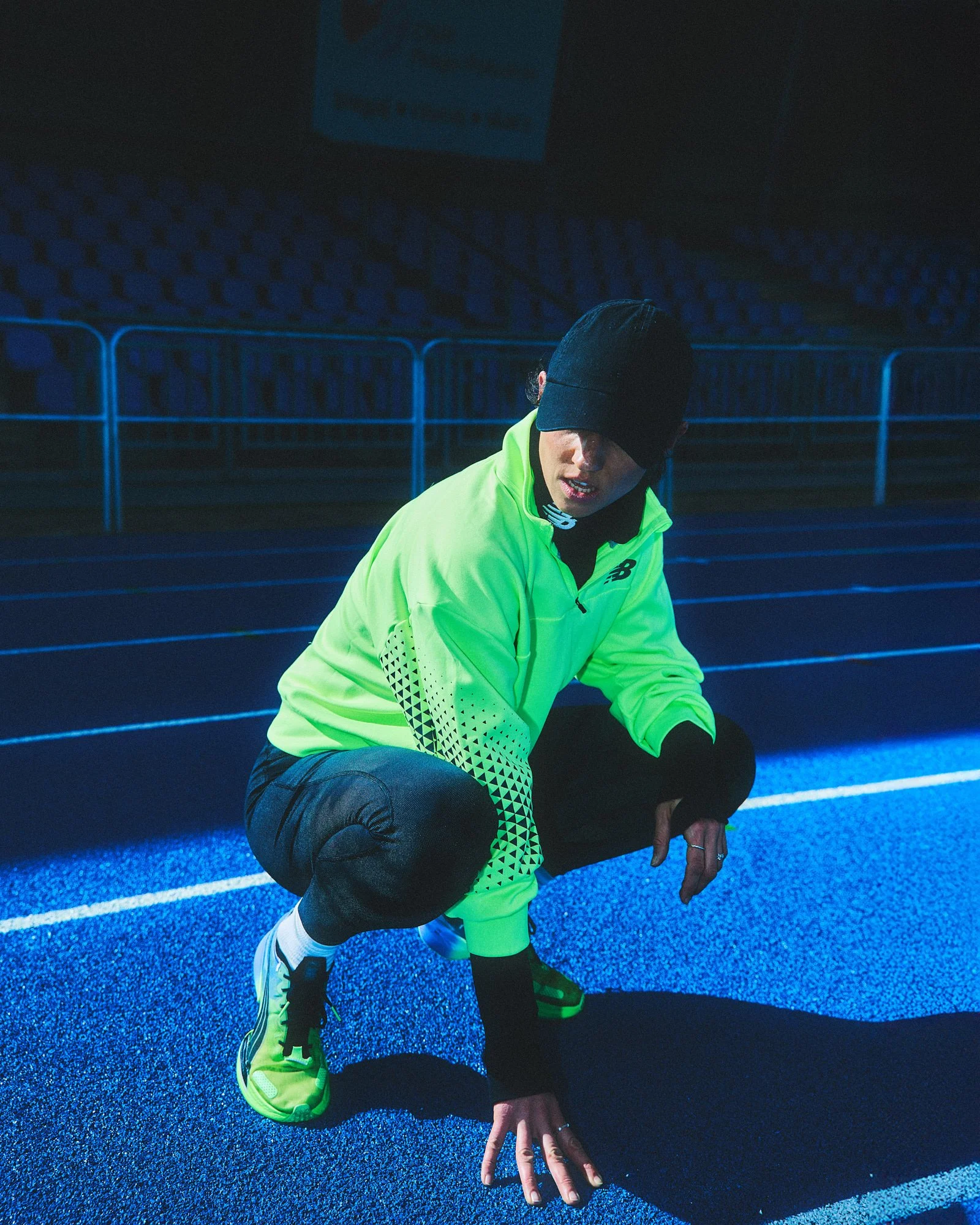 A person crouching on a blue running track at night, wearing a neon green sports jacket, black cap, black pants, and yellow running shoes.
