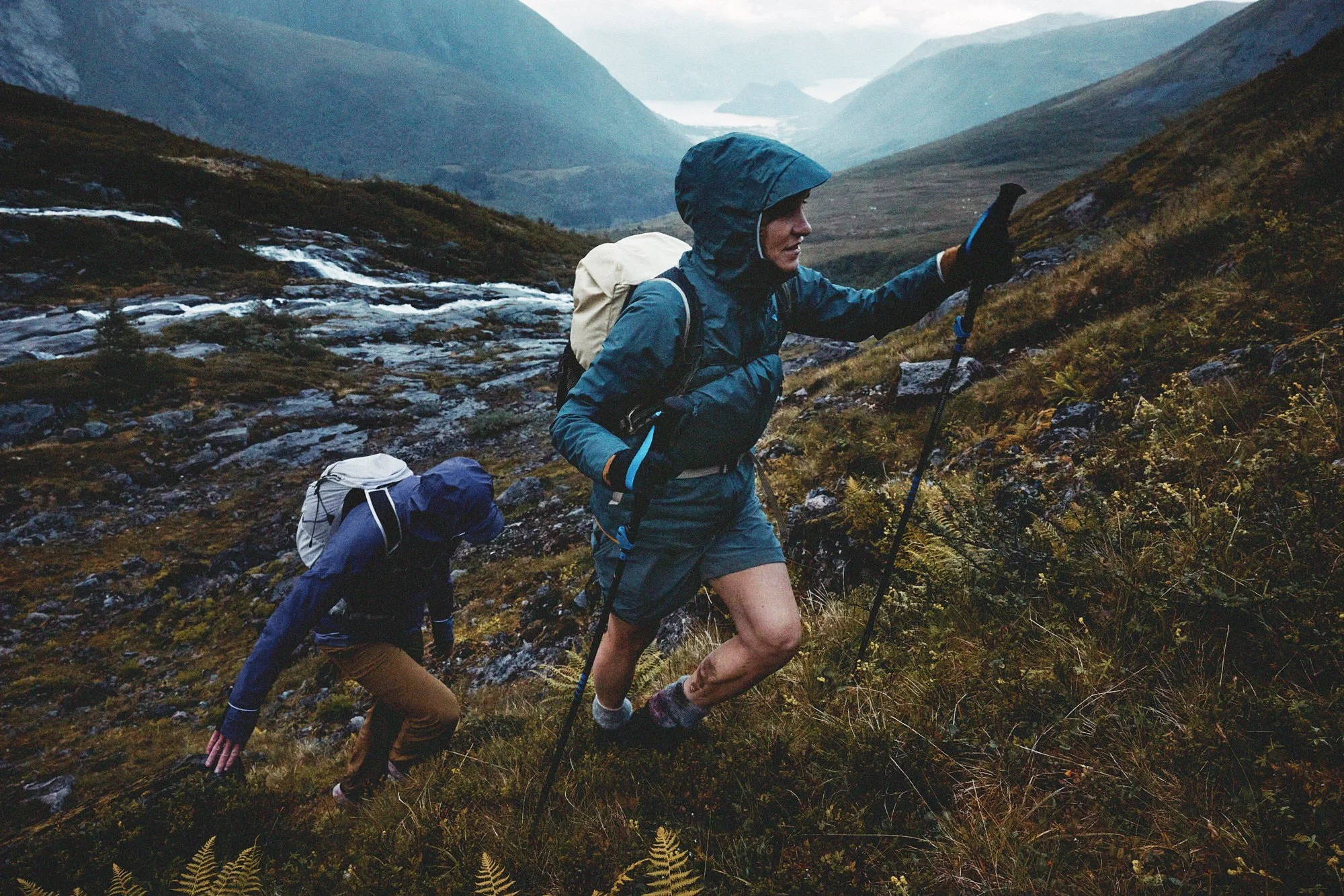 Two hikers climbing a grassy hillside with a mountain and water in the background, dressed in outdoor gear and carrying backpacks.