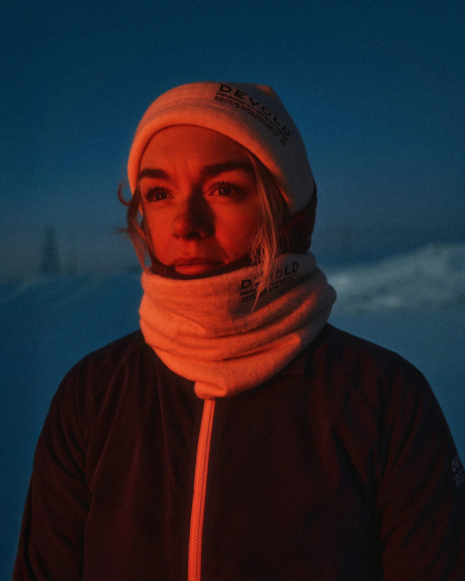 A woman wearing a white hat, neck gaiter, and dark jacket, standing outdoors in a snowy landscape at dusk or dawn with a blue sky in the background.
