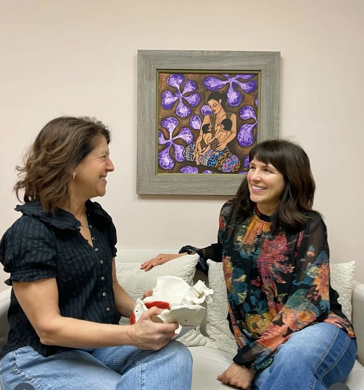 A physical therapist and physical therapy assistant are sitting on a white couch in a room with a beige wall and colorful artwork, smiling and engaging in conversation. One woman is holding a model of a pelvis.