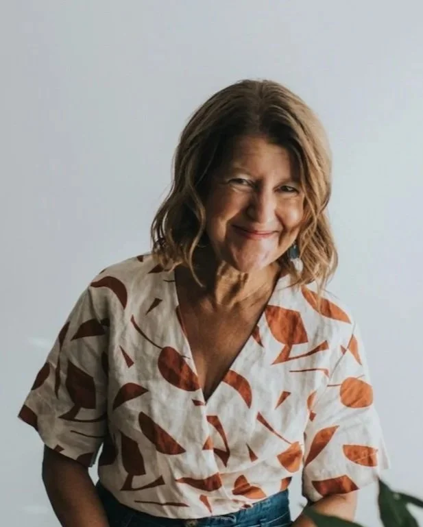 Smiling woman with shoulder-length brown hair wearing a cream and rust-colored patterned shirt, standing against a plain light background.
