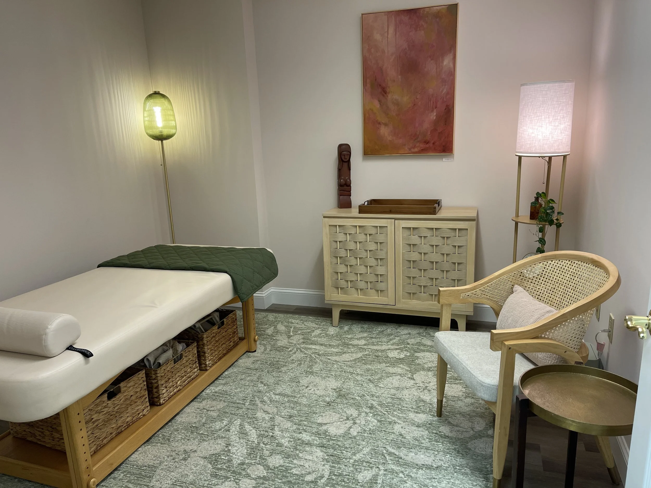 A room with a massage table, a wicker basket storage underneath, a green quilted blanket, and a pillow. There are two standing lamps, one with a green shade and another with a white shade. A cream-colored cabinet with a wooden tray on top is against the wall, and above it hangs an abstract pink and brown painting. There is a wooden chair with a woven back and a round coffee table with a gold finish.
