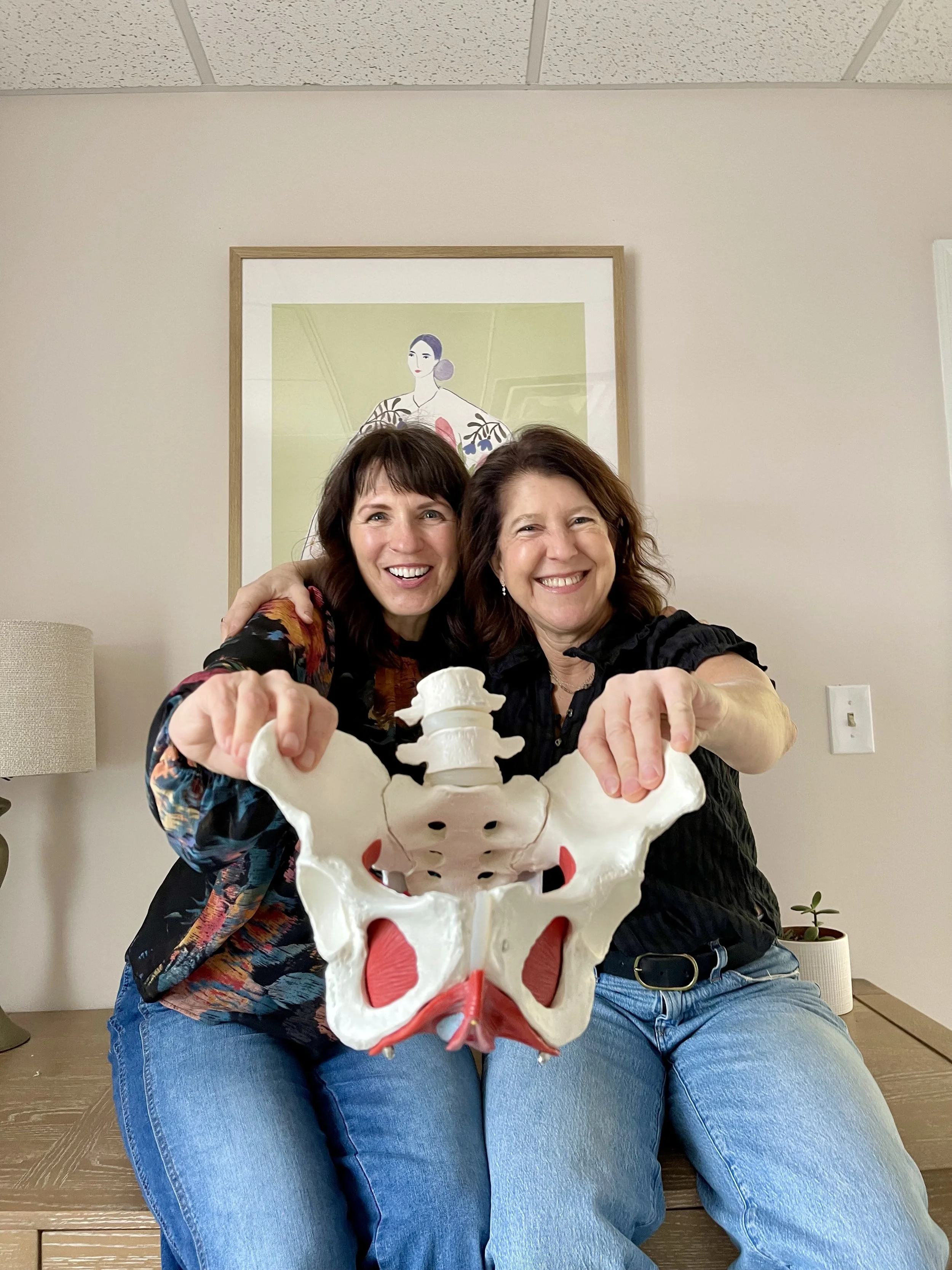 Two physical therapists sit on a desk, smiling and holding a large anatomical pelvis model facing the camera, with a framed artwork of a woman in traditional Japanese clothing hanging on the beige wall behind them.
