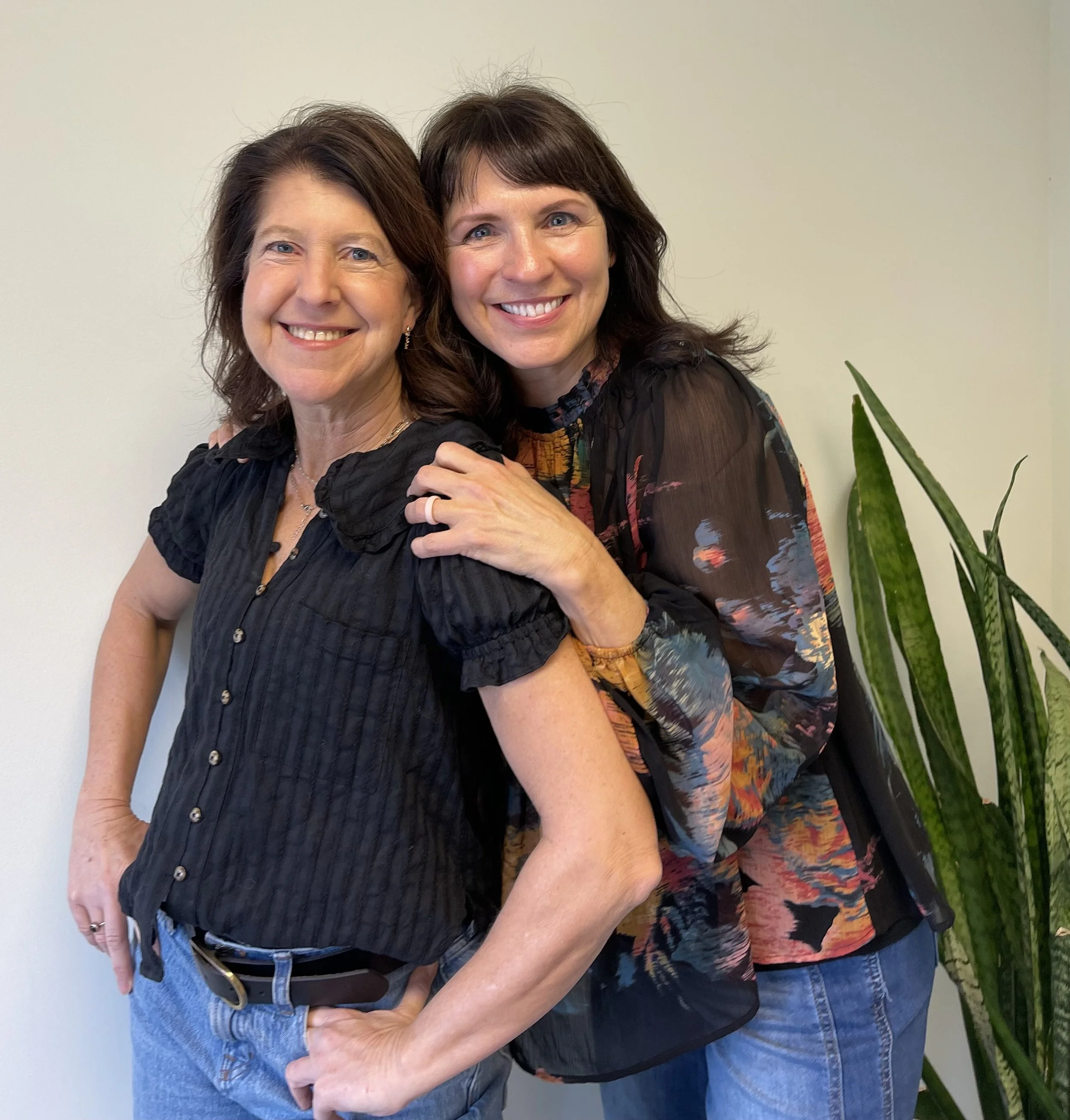 Two physical therapists, smiling, one with dark hair in a black blouse, the other with darker hair in a colorful blouse, with the woman in black having her hand on her hip and the other woman resting her hand on the shoulder of the woman.