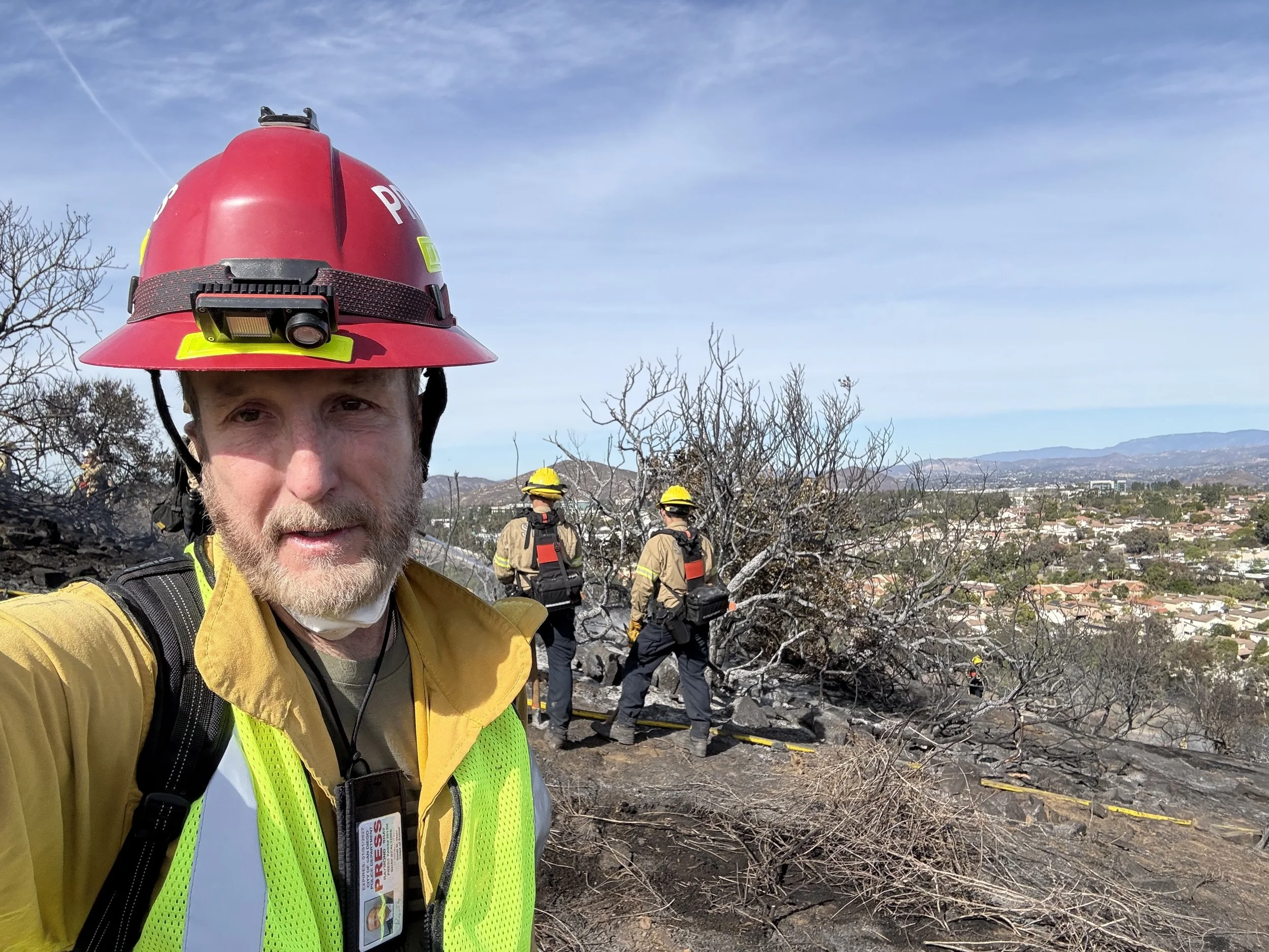 Firefighter taking a selfie at a burned area, wearing a red helmet with a headlamp and yellow protective clothing, with other firefighters in the background and a burnt landscape with sparse trees and a city in the distance under a clear blue sky.