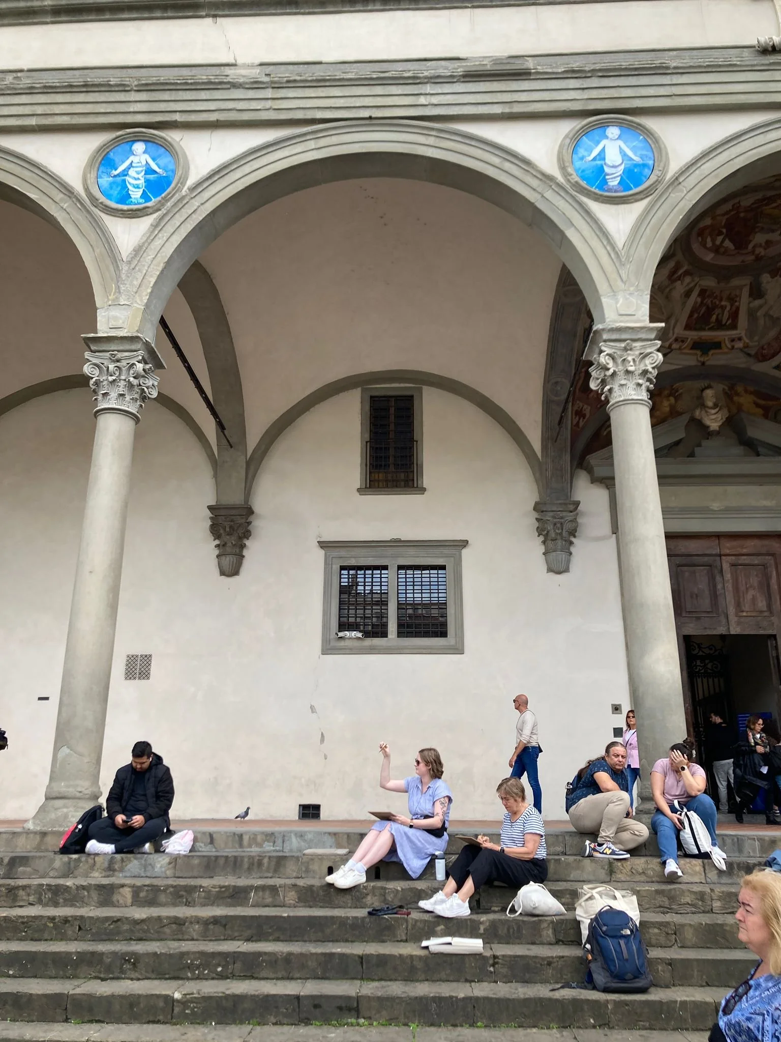People sitting on steps in front of a building with columns and arched windows, some using phones or tablets.