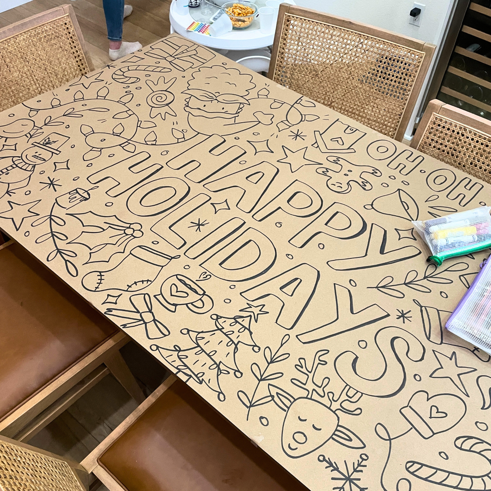 A large table with a holiday-themed coloring poster titled "Happy Holidays". The poster features outlined drawings of a reindeer, a snowman, a Christmas tree, a present, stars, snowflakes, holly, and other festive decorations. Coloring supplies are o