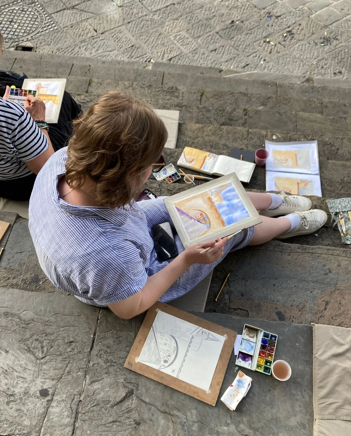 Person sitting on steps painting a watercolor landscape of a bridge over water with a blue sky, with art supplies around including paints, brushes, and paper.