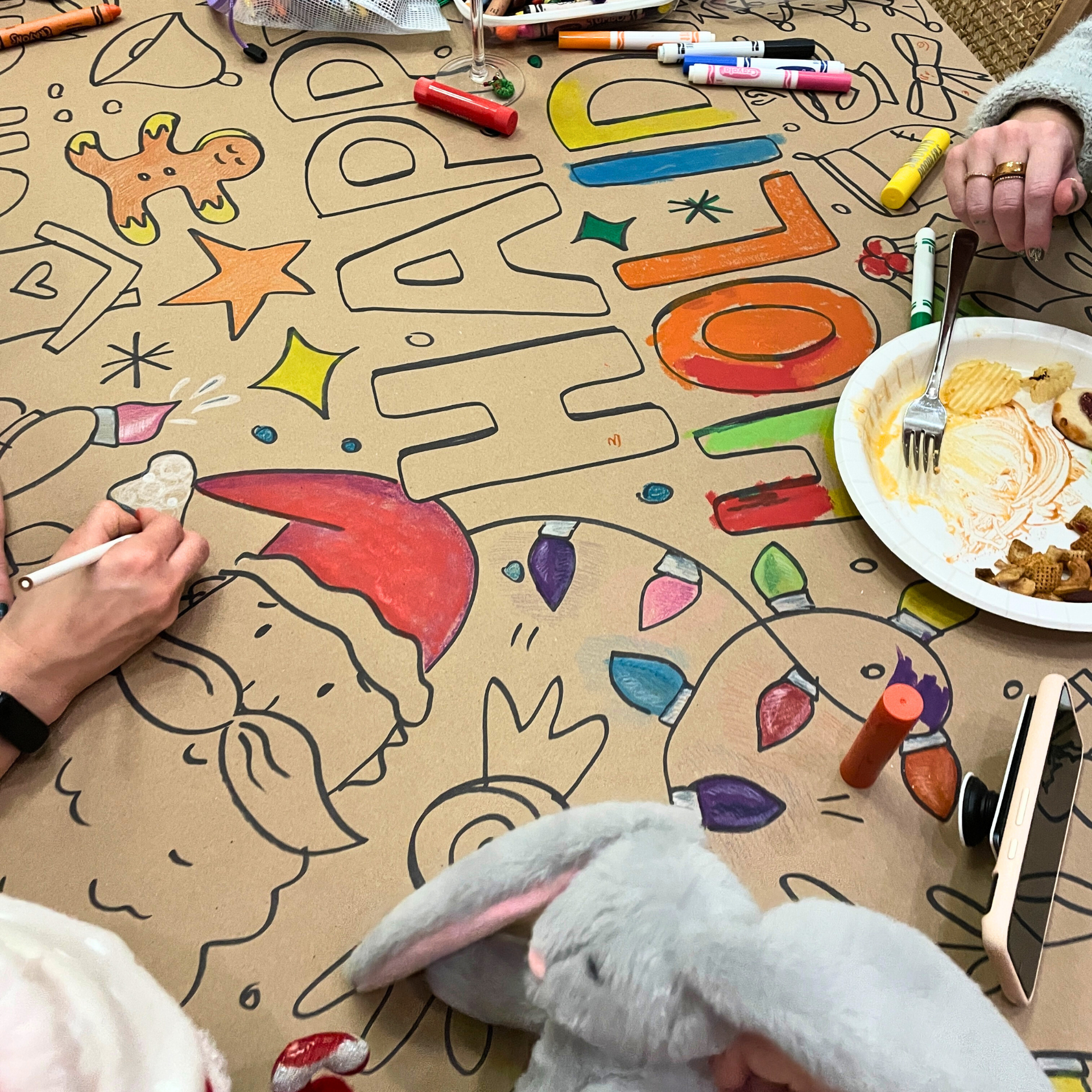 People decorating a large Christmas-themed poster with drawings of Santa Claus, Christmas lights, a reindeer, and holiday decorations, using markers and colored pens.