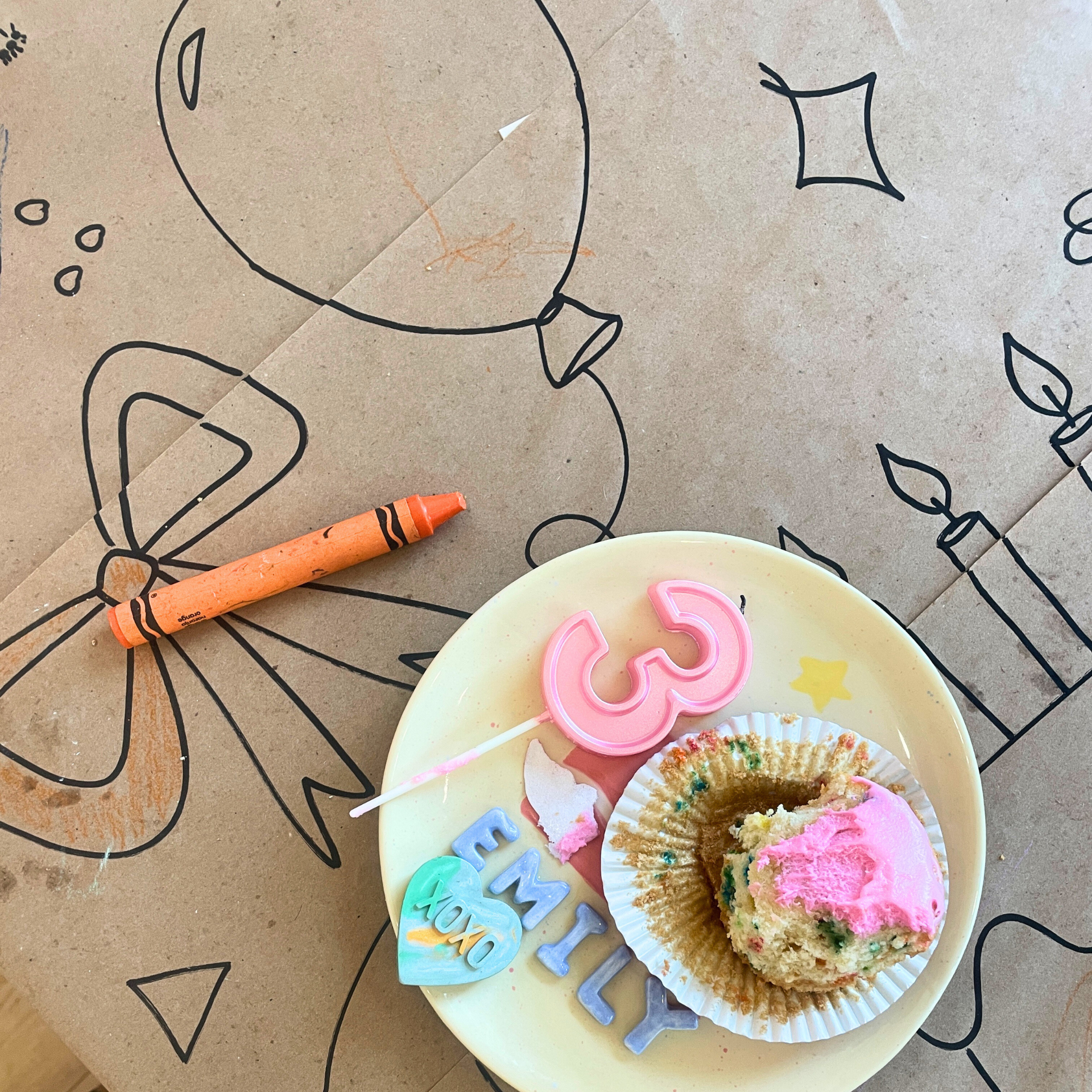 A birthday celebration with a cupcake on a yellow plate, decorated with a pink '25' candle, colorful 'EMILY' letters, and a blue heart-shaped decoration, set on a table with a drawing of a butterfly, candle, and star in the background.