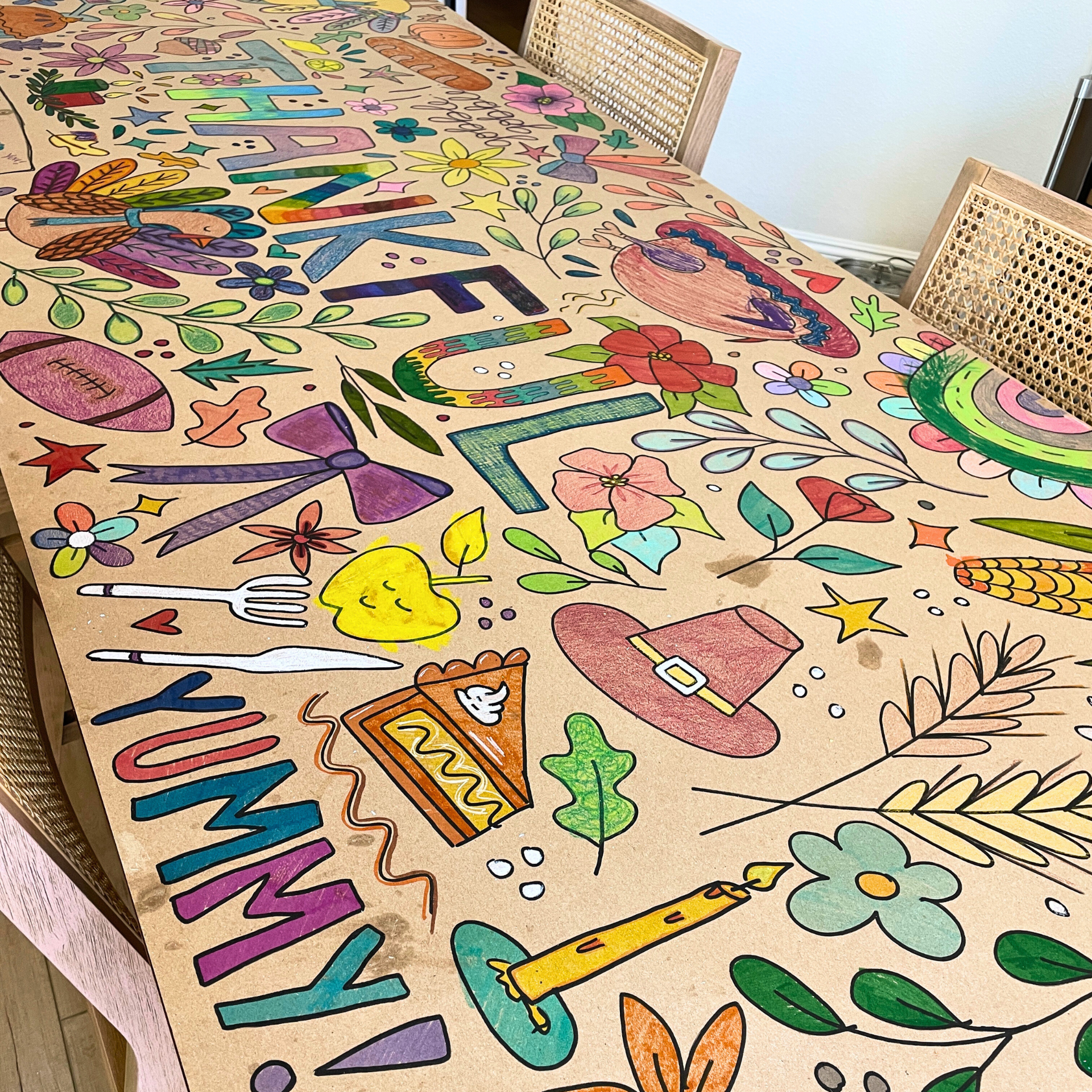 Colorful decorated table with drawings of flowers, leaves, a rainbow, a pilgrim hat, a turkey, a candle, and the word 'THANKFUL' written on the paper covering it.