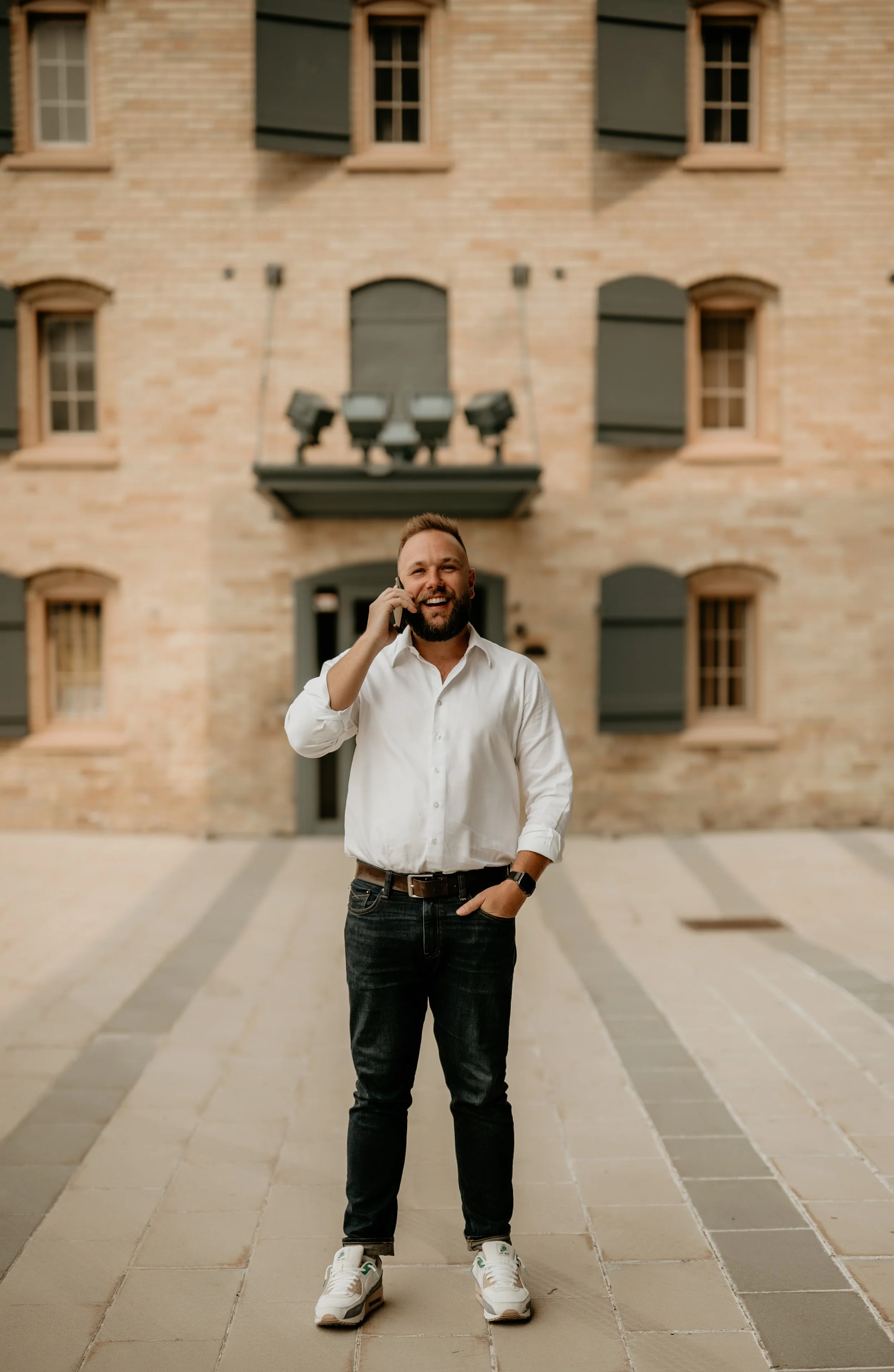 A man with a beard and short hair, wearing a white shirt, dark jeans, and sneakers, is talking on a cell phone outdoors in front of a brick building with multiple windows and a balcony.