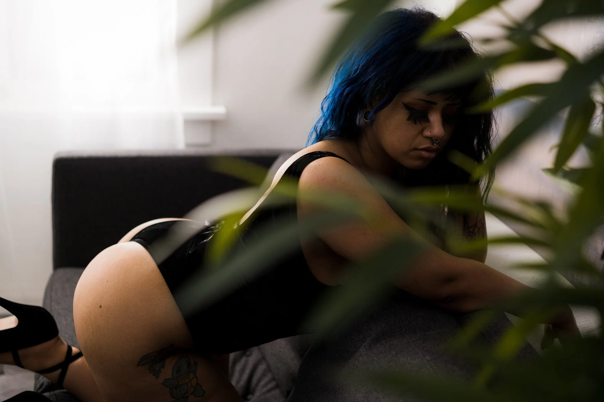 A woman with dark hair tinged with blue, wearing dark clothing, sitting with her head down in an indoor setting partially obscured by plant leaves.