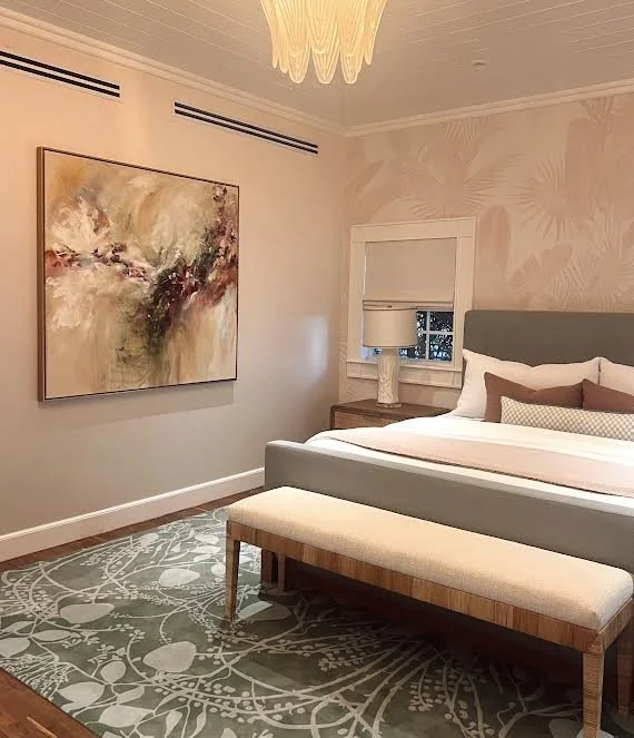 A bedroom featuring a bed with gray headboard, white and beige bedding, and decorative pillows. There is a wooden nightstand with a white lamp near a window with a white shade. An abstract wall art piece hangs above the bed, and a patterned area rug covers part of the floor. The room has soft pastel-colored wallpaper with a leaf design.