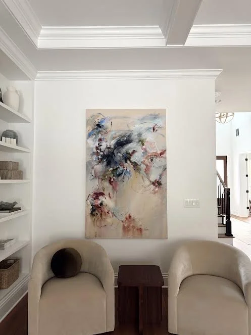 Living room with a white wall, abstract painting, two beige chairs, and a small brown side table.
