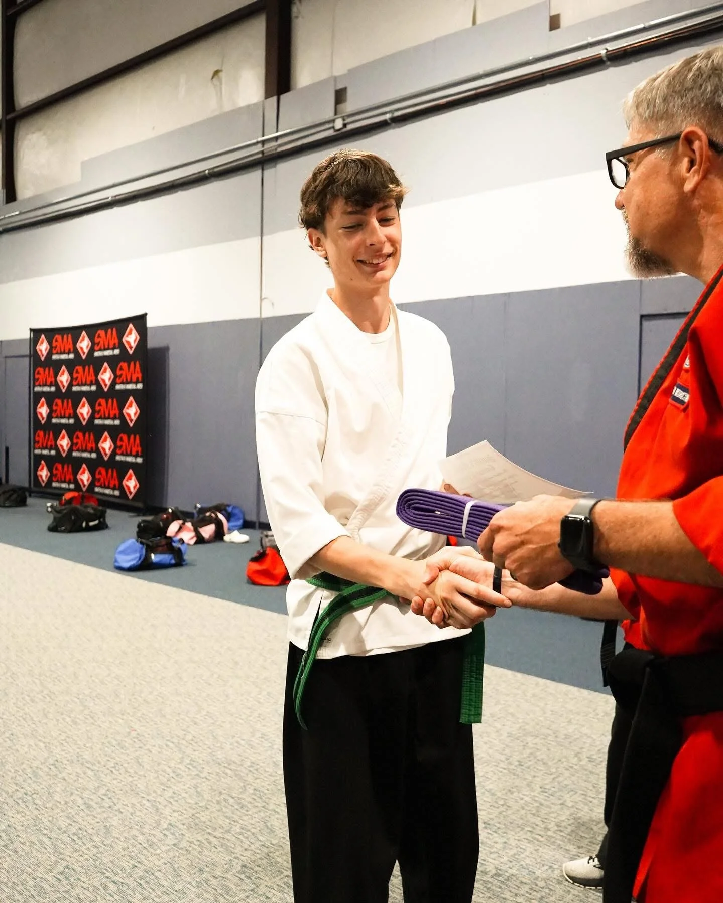A young man in a white martial arts gi shaking hands with an instructor during a martial arts award ceremony, with martial arts gear and SMA banners in the background.