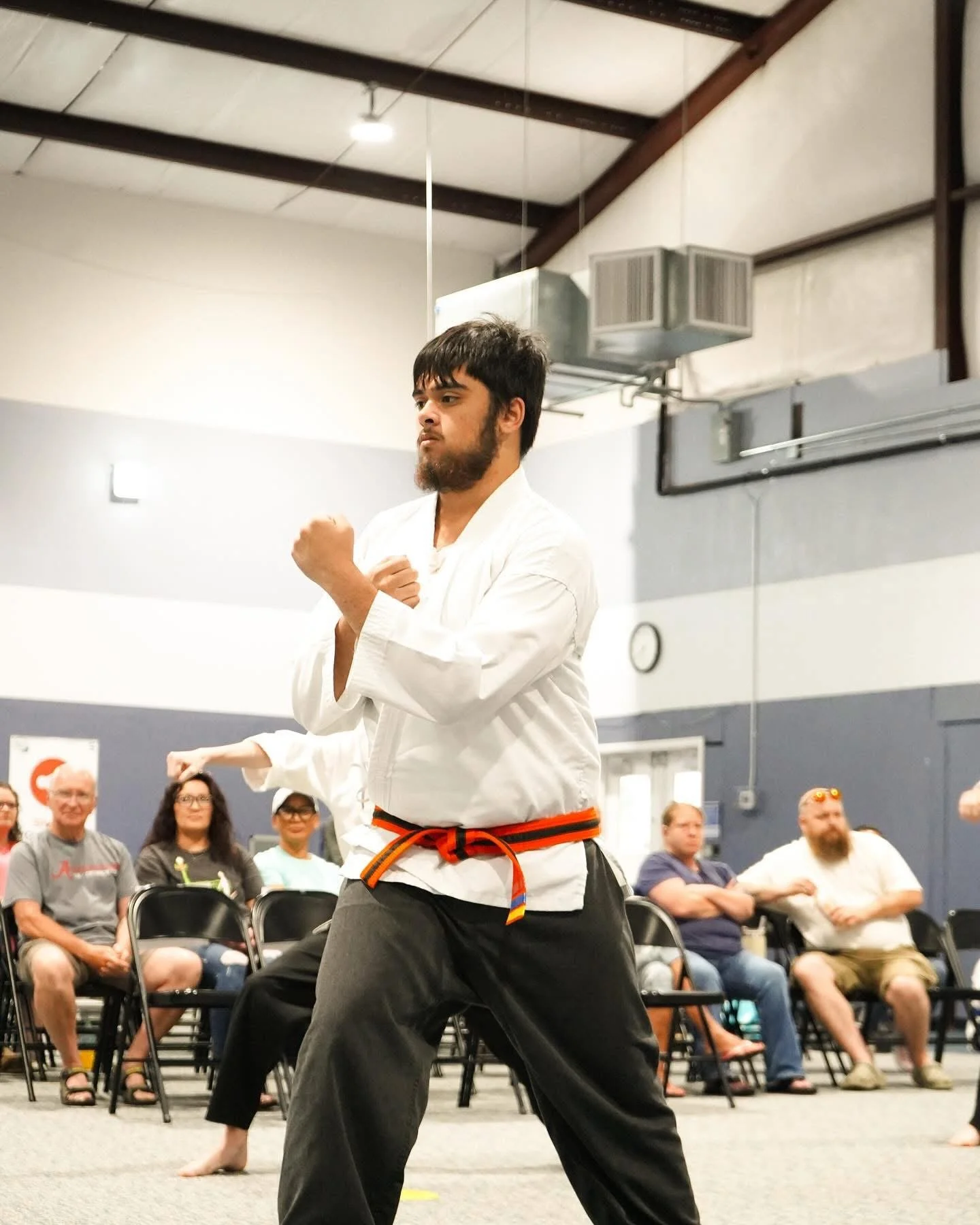 Martial artist in a white gi and black belt demonstrating a stance during a class with seated audience watching.