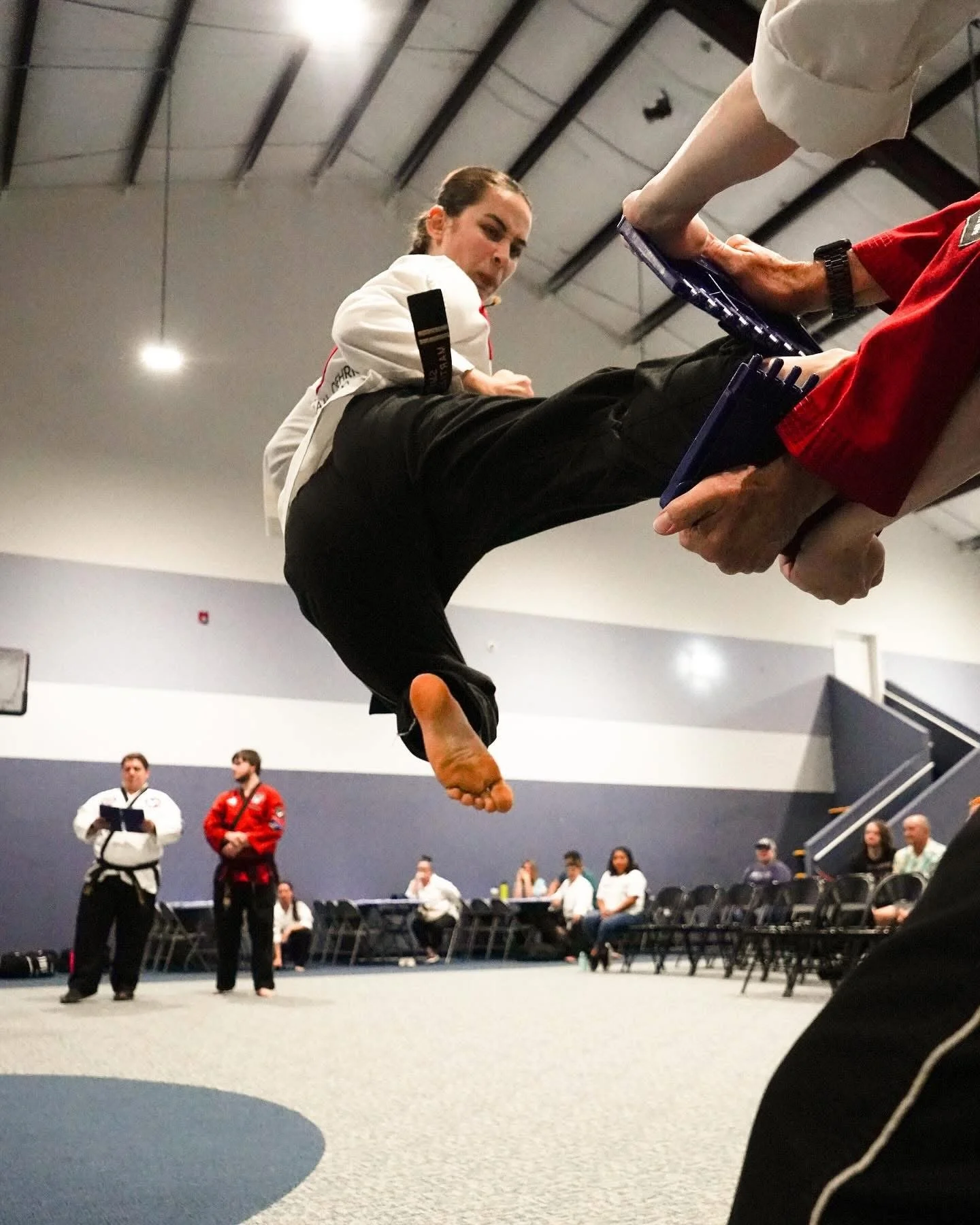 Female martial artist in a white gi executing a flying kick during a martial arts demonstration or competition in an indoor gymnasium, with judges and audience watching.