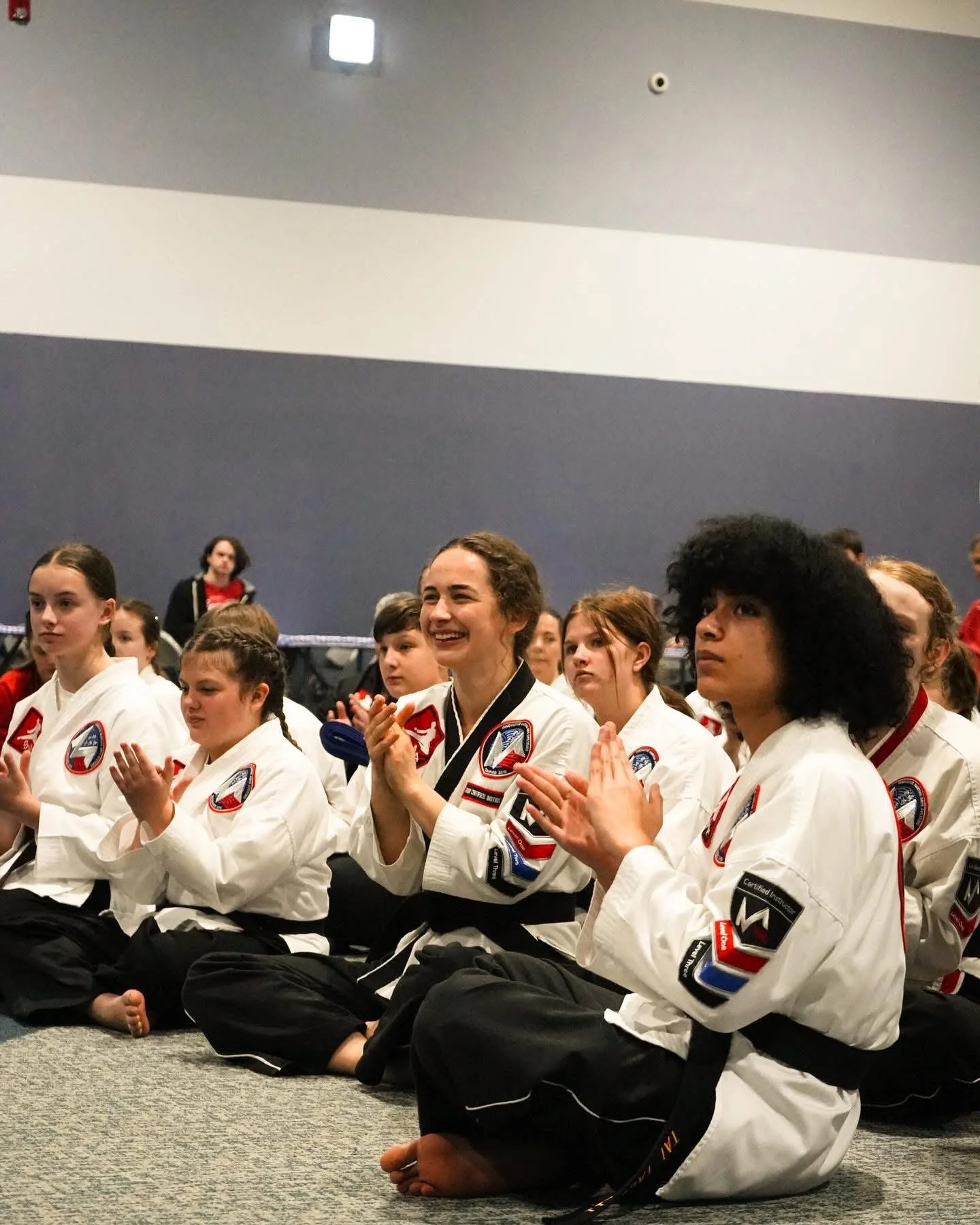 Kids in martial arts uniforms sitting on the floor, clapping and smiling in a dojo or martial arts training center.